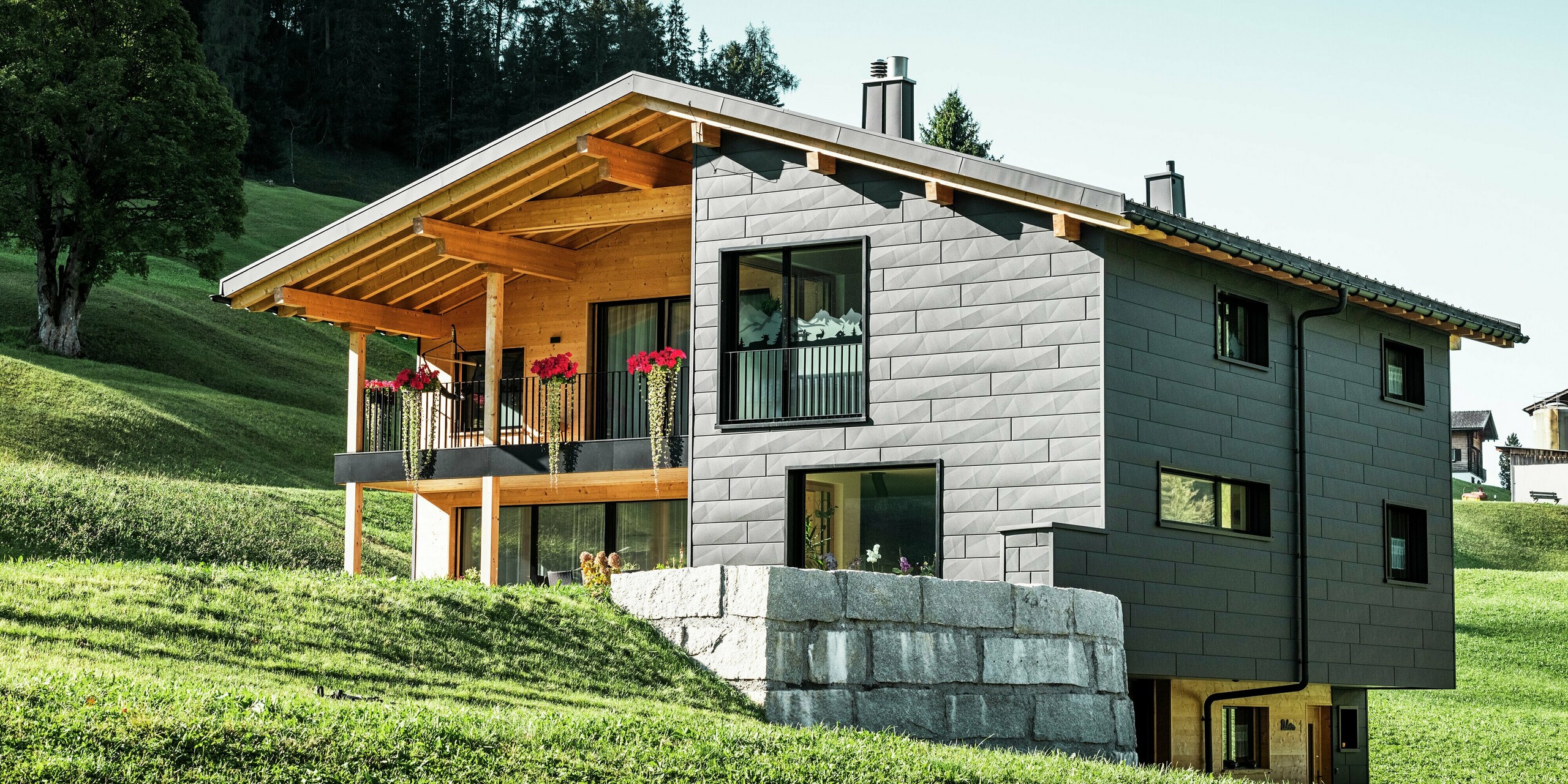 Dieses moderne Einfamilienhaus in Ascharina, Schweiz, fügt sich harmonisch in die grüne Alpenlandschaft ein. Das Gebäude ist komplett mit den langlebigen und witterungsbeständigen PREFA Siding.X Fassadenpaneelen in P.10 Schwarz verkleidet, die dem Bauwerk ein einzigartiges Erscheinungsbild verleihen. Der Holzbalkon ergänzt die dunkle Aluminiumhülle perfekt und setzt einen natürlichen Akzent.