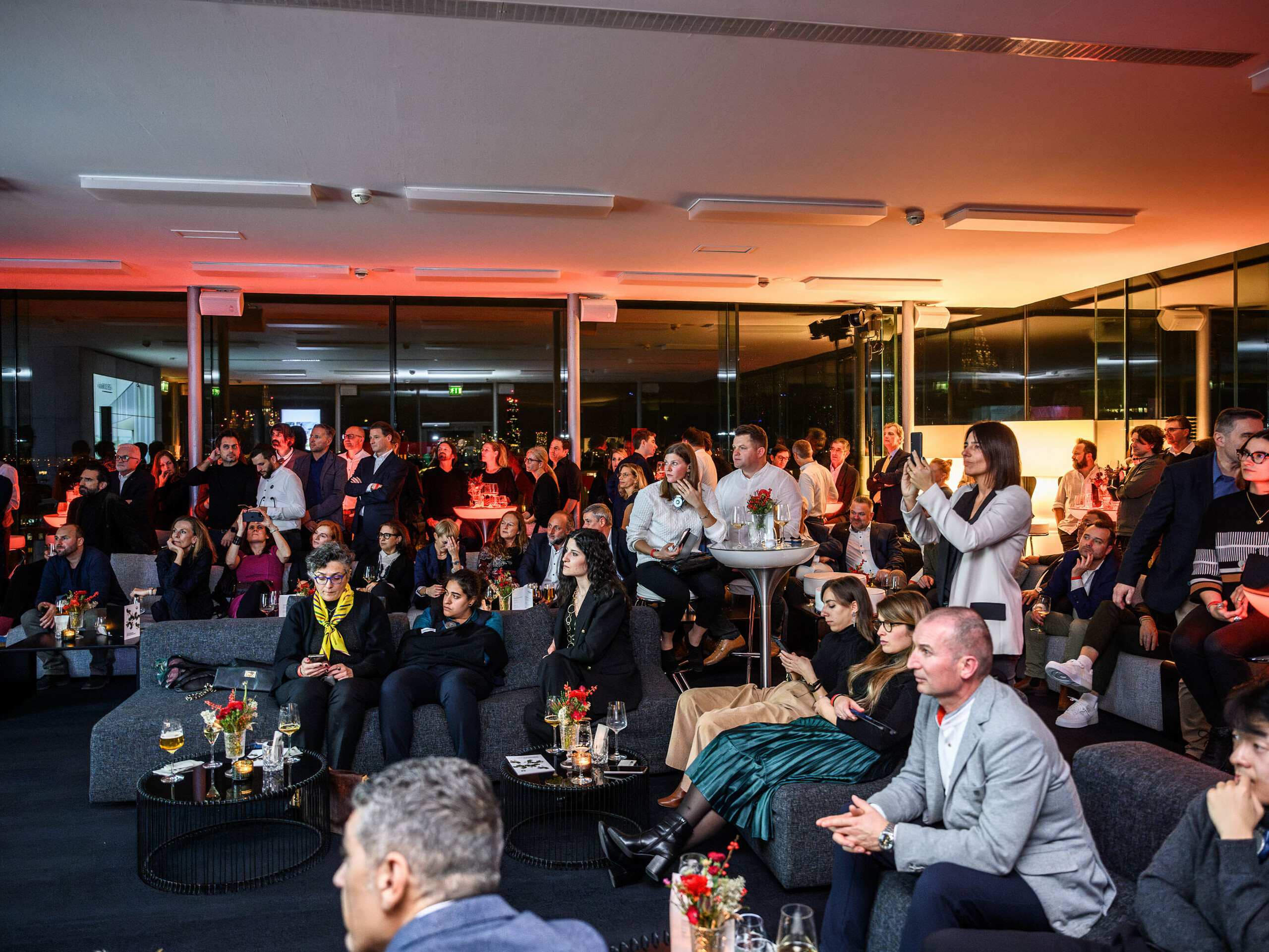 Die Besucher:innen auf einen Blick bei der PREFA Prefarenzen Präsentation für das Jahr 2024, bei der es um außergewöhnliche Architektur geht. Einige sitzen auf Sofas oder stehen mit Getränken und kleinen Tischen vor sich.