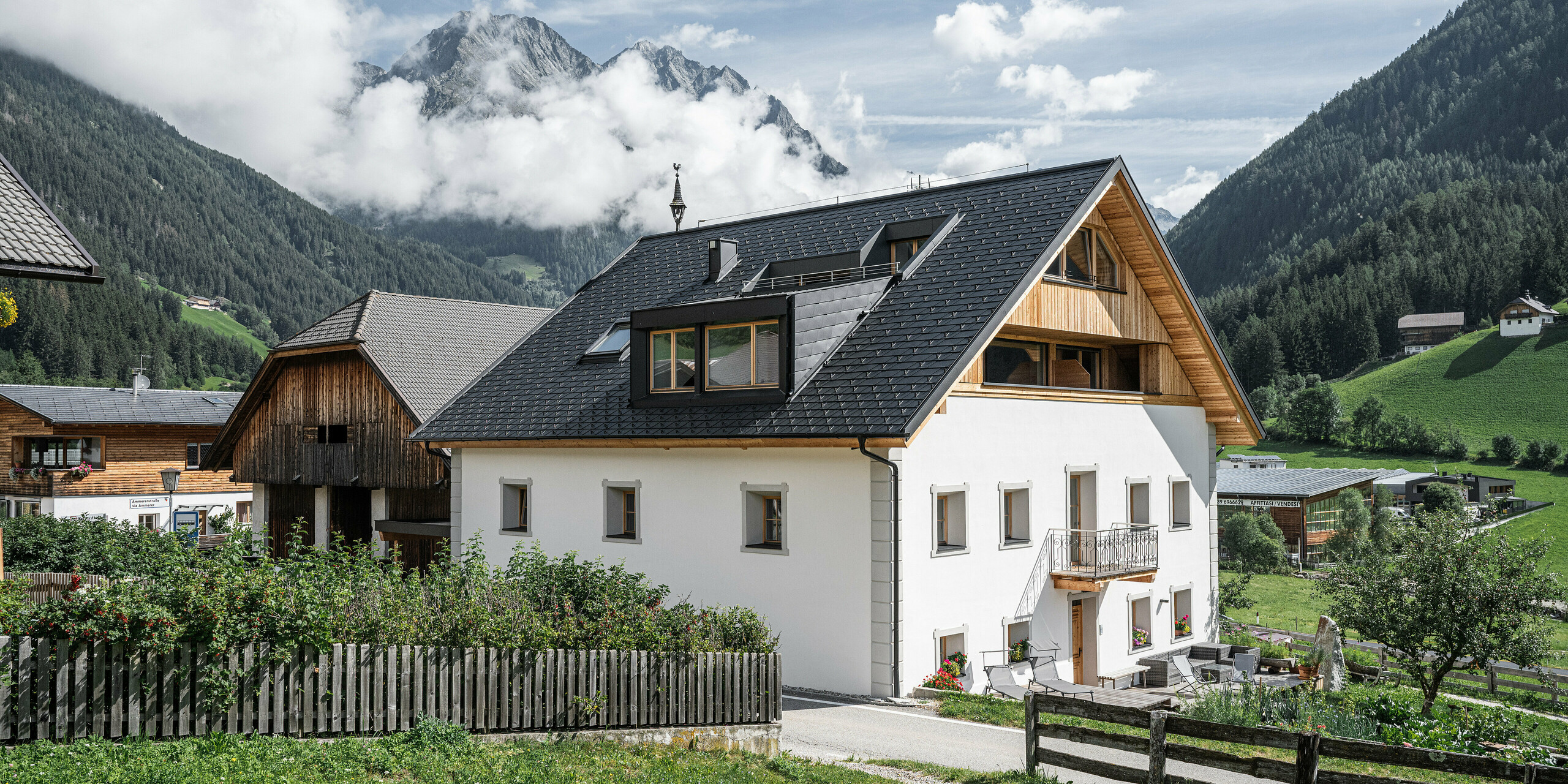 Gesamtansicht der Ferienwohnungen Ausserwegerhof im idyllischen Antholzertal, Südtirol, eingedeckt mit PREFA DS.19 Dachschindeln in Anthrazit. Das traditionelle Gebäude verbindet sich mit modernen Elementen und bietet einen malerischen Blick auf die majestätische Bergkulisse, die geheimnisvoll von Wolken umgeben ist. Der Ausserwegerhof in der Mitte des Bildes ist umgeben von klassischen Bauernhöfen und grünen Wiesen.