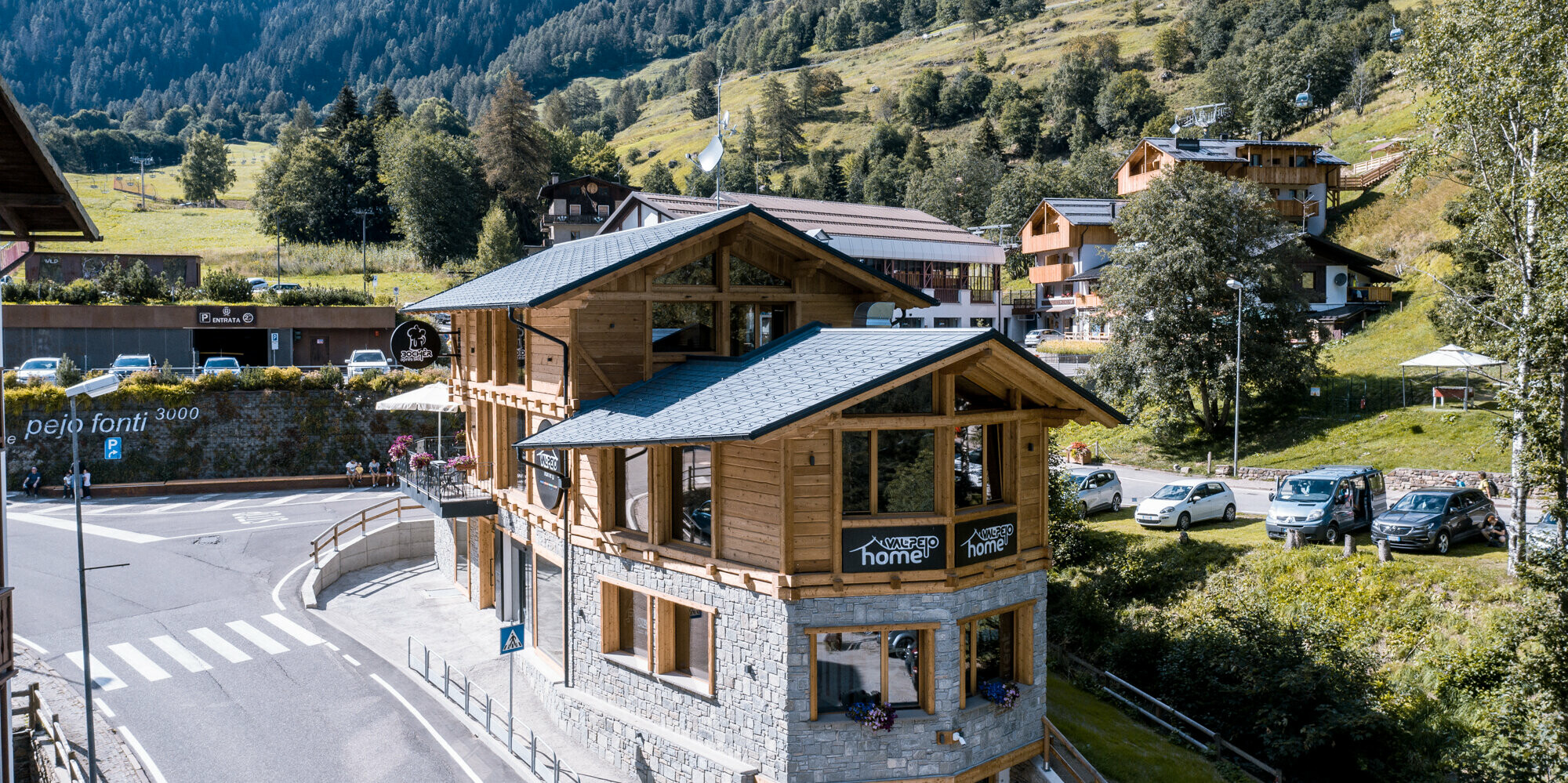 Ansicht einer modernen Après-Ski-Bar in traditioneller Holzarchitektur mit steingrauem PREFA Dachschindeln DS.19, aus Aluminium integriert in die idyllische Berglandschaft mit üppigem Grün und Blick auf die Alpen.