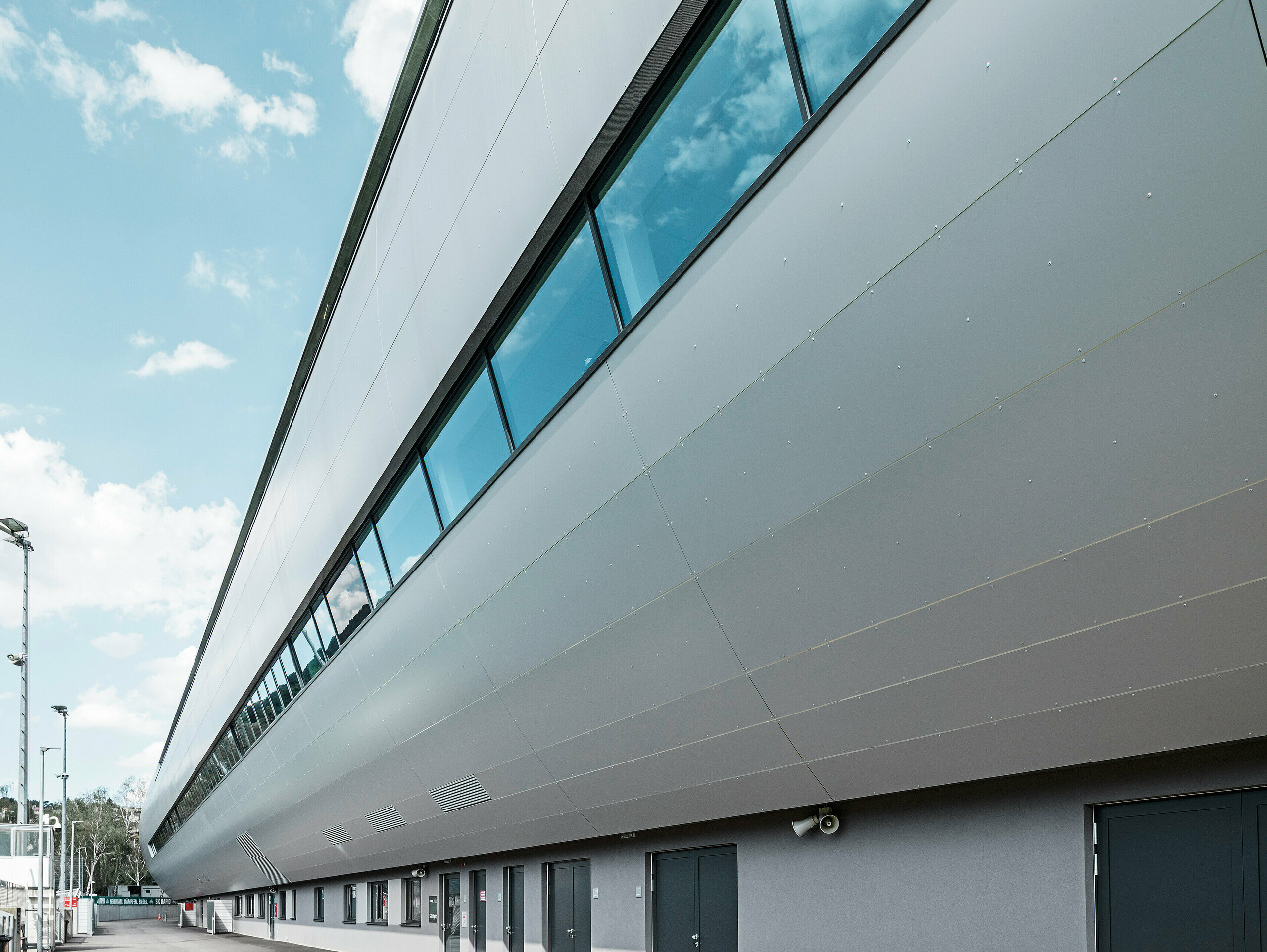 Seitenansicht des Allianz Stadions von SK Rapid Wien, verkleidet mit PREFA Aluminium Verbundplatten in Silbermetallic. Die Fassade zeigt die beeindruckende Länge und klare Linienführung des Bauwerks, während die großflächigen Fenster den Blick zum Trainingsplatz rahmen. In den Fensterscheiben reflektiert der blaue Himmel. Die hochwertige Metallverkleidung verkörpert Beständigkeit und moderne Architektur.