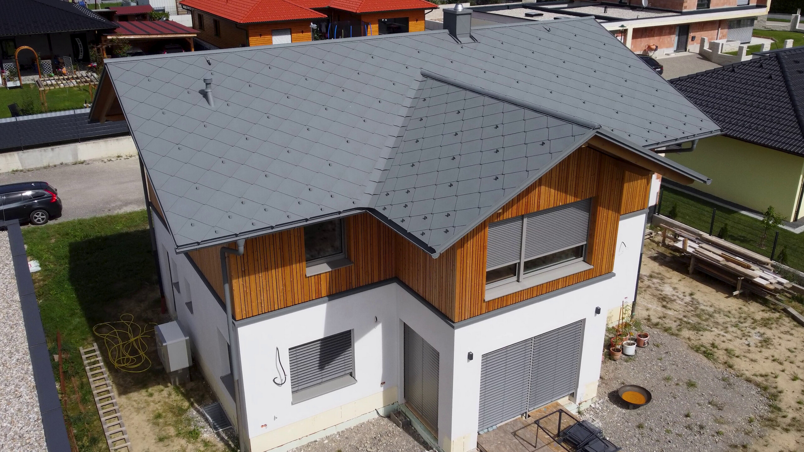 Ein Einfamilienhaus mit einem PREFA Dach in Rautenform, ausgeführt in hellgrau. Die moderne Architektur kombiniert das metallische Dach mit einer Fassade aus weißen und Holzelementen. Sonnenschutzlamellen und Rollläden an den Fenstern ergänzen das Design, während die umliegende Landschaft aus gepflasterten Wegen und sorgfältig gepflegten Grünflächen das Bild abrunden.