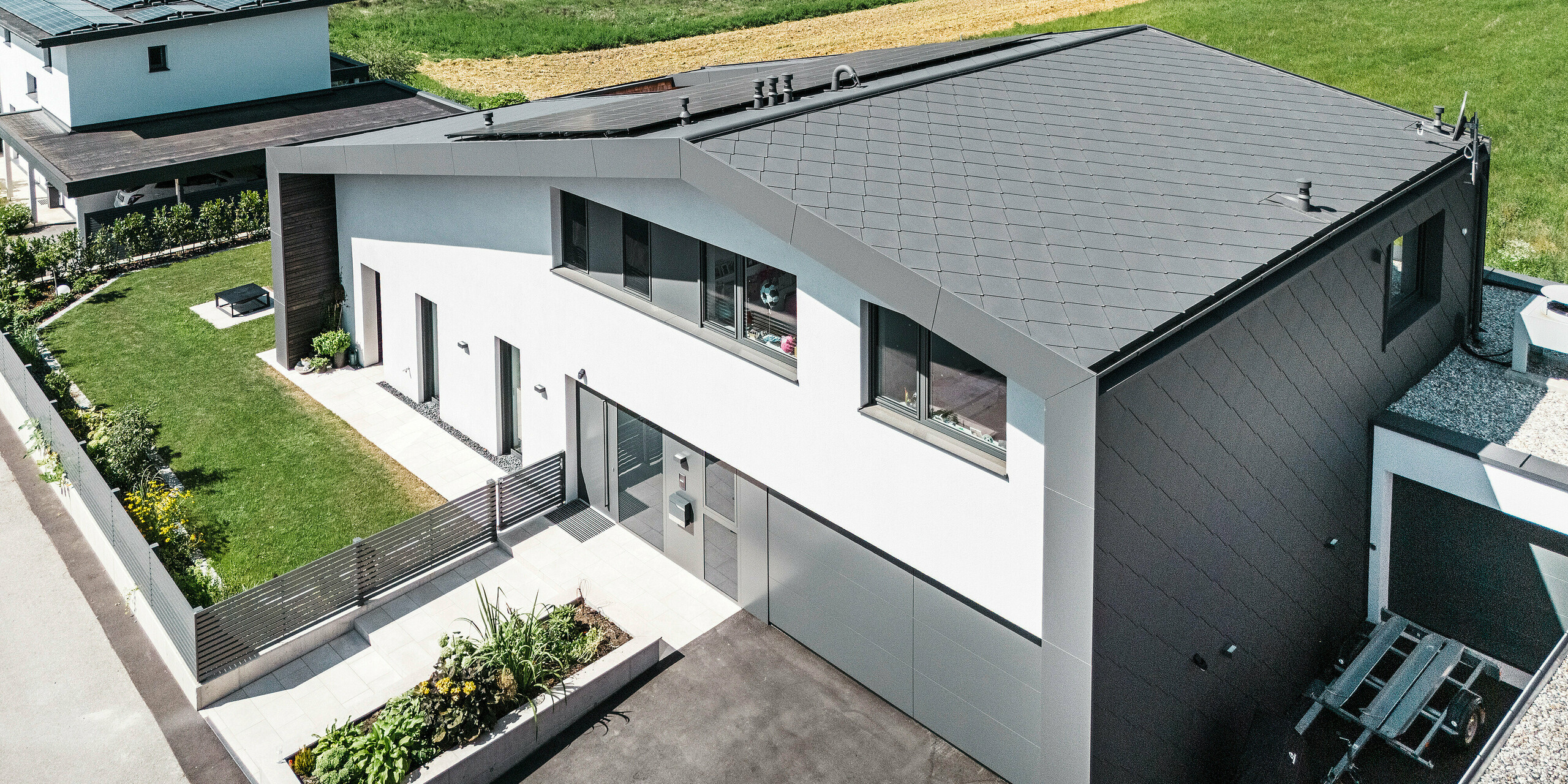 Luftaufnahme eines modernen Einfamilienhauses in Aschach mit zeitloser Architektur. Fassade und Dach sind mit dunkelgrauen PREFA Aluminium-Rauten 44 × 44 eingedeckt. Die langlebige und witterungsbeständige Hülle verleiht dem Gebäude ein elegantes Aussehen und bietet optimalen Schutz vor Umwelteinflüssen. Vor dem Haus befindet sich ein gepflegter Vorgarten mit einer saftig grünen Wiese. Rechts vom Haus liegt ein kleines Nebengebäude und neben der Eingangstür befindet sich eine großzügige Garage mit asphaltierter Zufahrt.