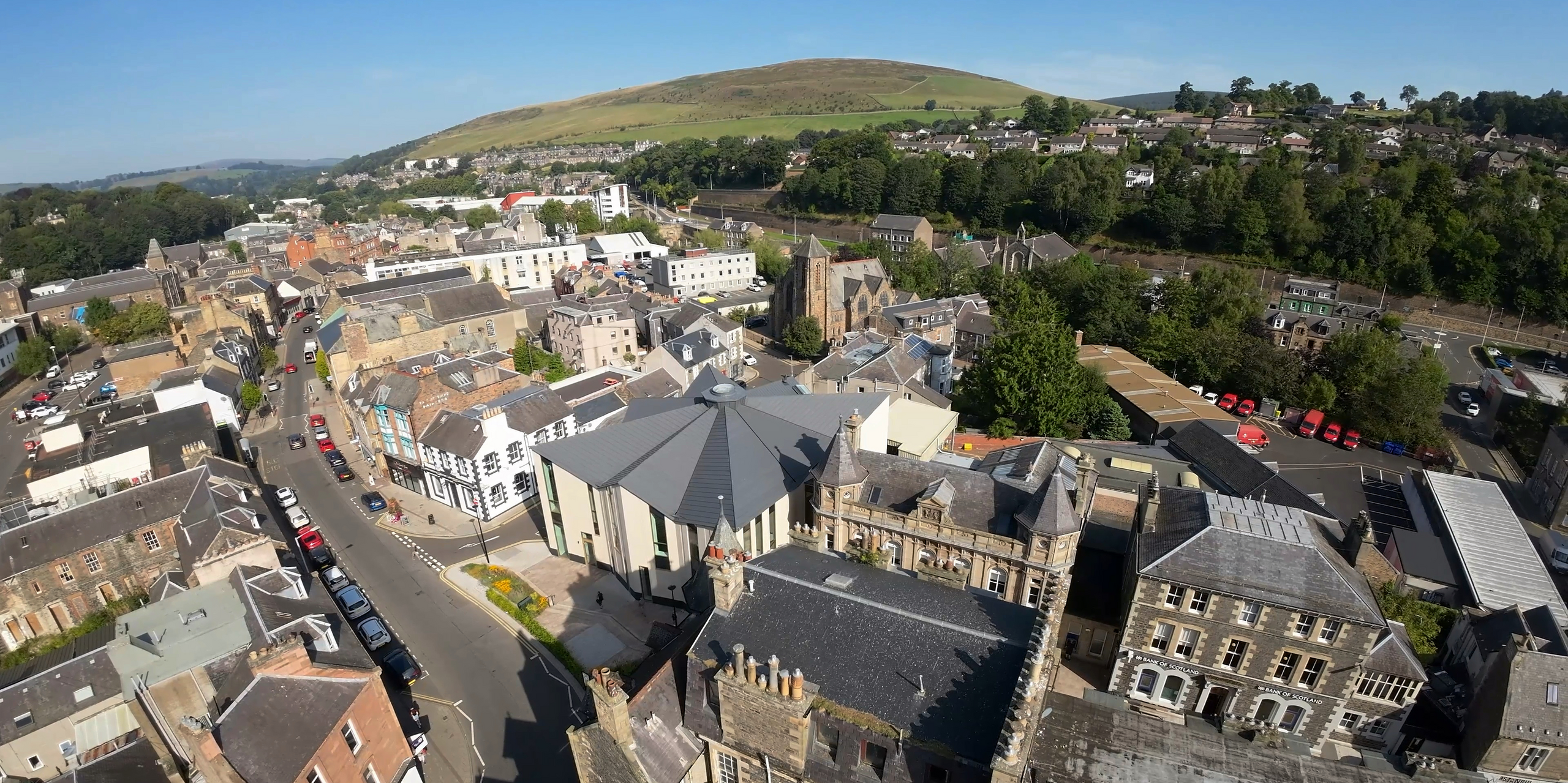 Weitwinkel-Drohnenaufnahme der Galerie bzw. Besucherzentrums 'The Great Tapestry of Scotland' in Galashiels, umrahmt von der malerischen Hügellandschaft Schottlands. Das markante Gebäude sticht mit seinem PREFALZ Dachsystem in P.10 Zinkgrau hervor, das sich elegant über die einzigartige Struktur wölbt. Die außergewöhnlichen Linien des Daches heben sich deutlich von den umliegenden traditionellen Gebäuden ab und zeigen, wie PREFA Aluminiumprodukte moderne Architektur mit dem kulturellen Erbe der Stadt verbindet.