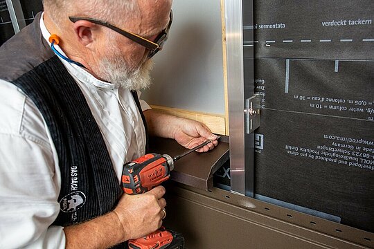 Der PREFA Techniker steckt die selbst gefalzte Fensterbank unterseitig am Fenster in die Profilnut und befestigt diese mithilfe eines Bohrer.