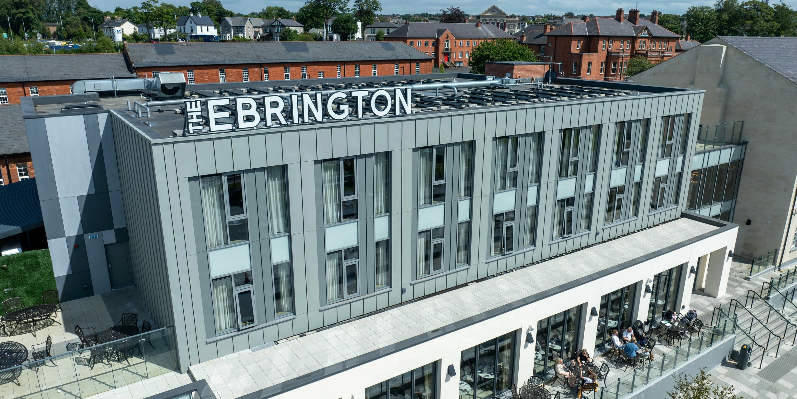 Das Ebrington Hotel im Zentrum von Londonderry, Nordirland, beeindruckt mit seiner modernen PREFALZ Aluminiumverkleidung in den Farben P.10 Hellgrau und P.10 Zinkgrau. Diese hochwertige, widerstandsfähige Blechfassade verleiht dem Gebäude eine elegante und robuste Optik. Die Gebäudehülle betont die zeitgemäße Architektur des Hotels, während die Terrasse im Vordergrund einen einladenden Platz für Gäste bietet. Im Hintergrund sind historische Gebäude zu sehen, die einen interessanten Kontrast zur modernen Gestaltung des Ebrington Hotels bilden.