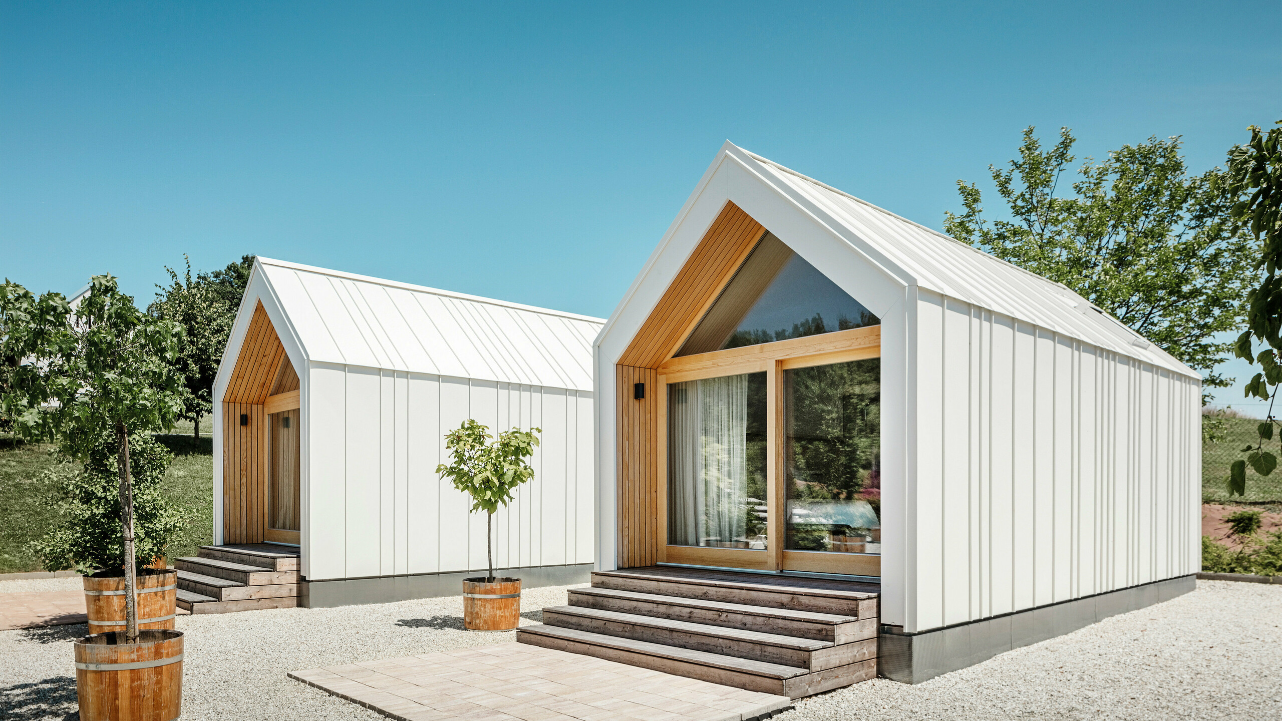 Zwei weiße Tiny Häuser der Ferienidylle 'Pri Momi' in Kančevci, Slowenien, gestaltet mit PREFALZ Dach- und Fassadenverkleidung in P.10 Reinweiß und naturbelassenem Holz. Die minimalistische Architektur der Gebäude wird durch den eleganten Materialmix aus Aluminium und Holz unterstrichen. Die großen Glasfronten an den Eingängen sowie die Holzstufen und umliegenden Bäume schaffen eine einladende und harmonische Atmosphäre. Die exklusive Unterkunft liegt inmitten einer malerischen Landschaft und wird speziell für Feiern genutzt.