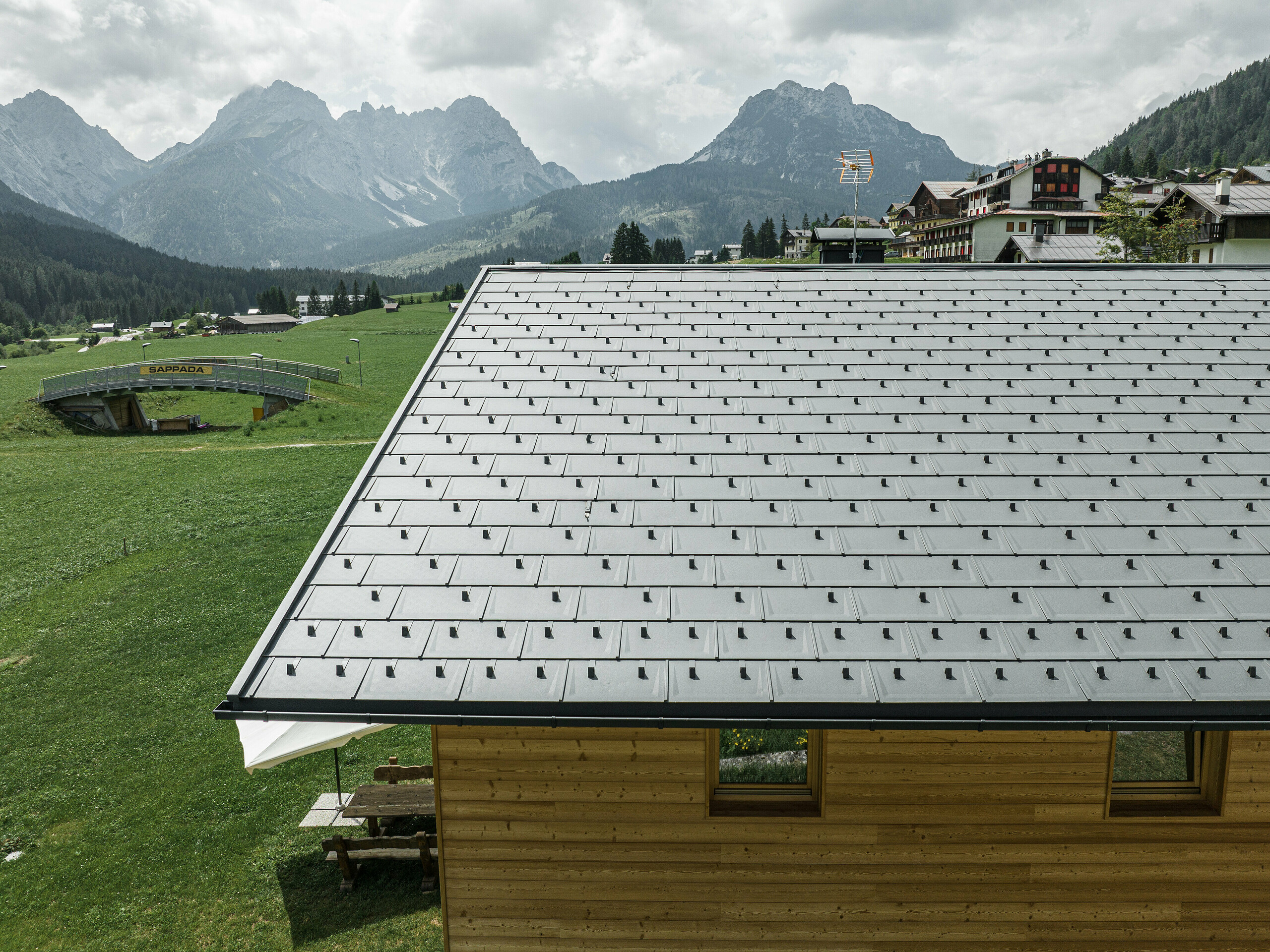 Dachansicht eines Einfamilienhauses in Sappada, Italien, umgeben von der majestätischen alpinen Kulisse. Das Haus ist mit PREFA Dachplatten R.16 in P.10 Schwarz ausgestattet, die ein modernes, lineares Muster bilden und einen ansprechenden Kontrast zur natürlichen Holzfassade darstellen. Diese hochwertigen, widerstandsfähigen Aluminiumdachplatten bieten einen langfristigen Schutz und unterstreichen das Engagement von PREFA für Ästhetik und Funktionalität.