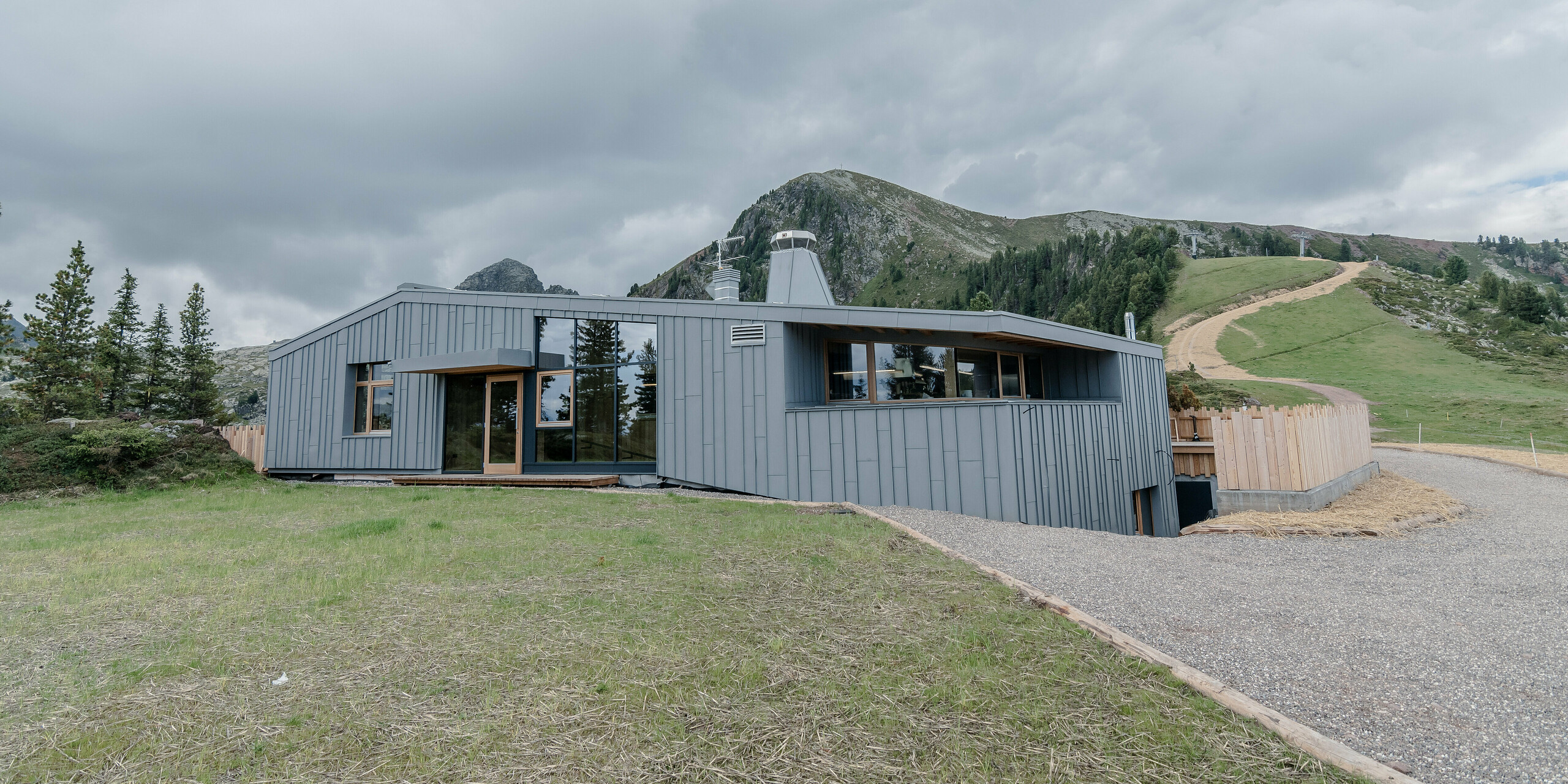 Das Ristorante Busabelle, eingebettet in eine idyllische Berglandschaft, ist von einer Winkelstehfalz Spiegeldeckung aus hellgrauem PREFALZ ummantelt, die die moderne Architektur hervorhebt und eine harmonische Verbindung zur natürlichen Umgebung schafft. Der Eingangsbereich ist mit großen Glasflächen versehen. Im Hintergrund sind Masten für eine Sesselliftanlage zu erkennen. 