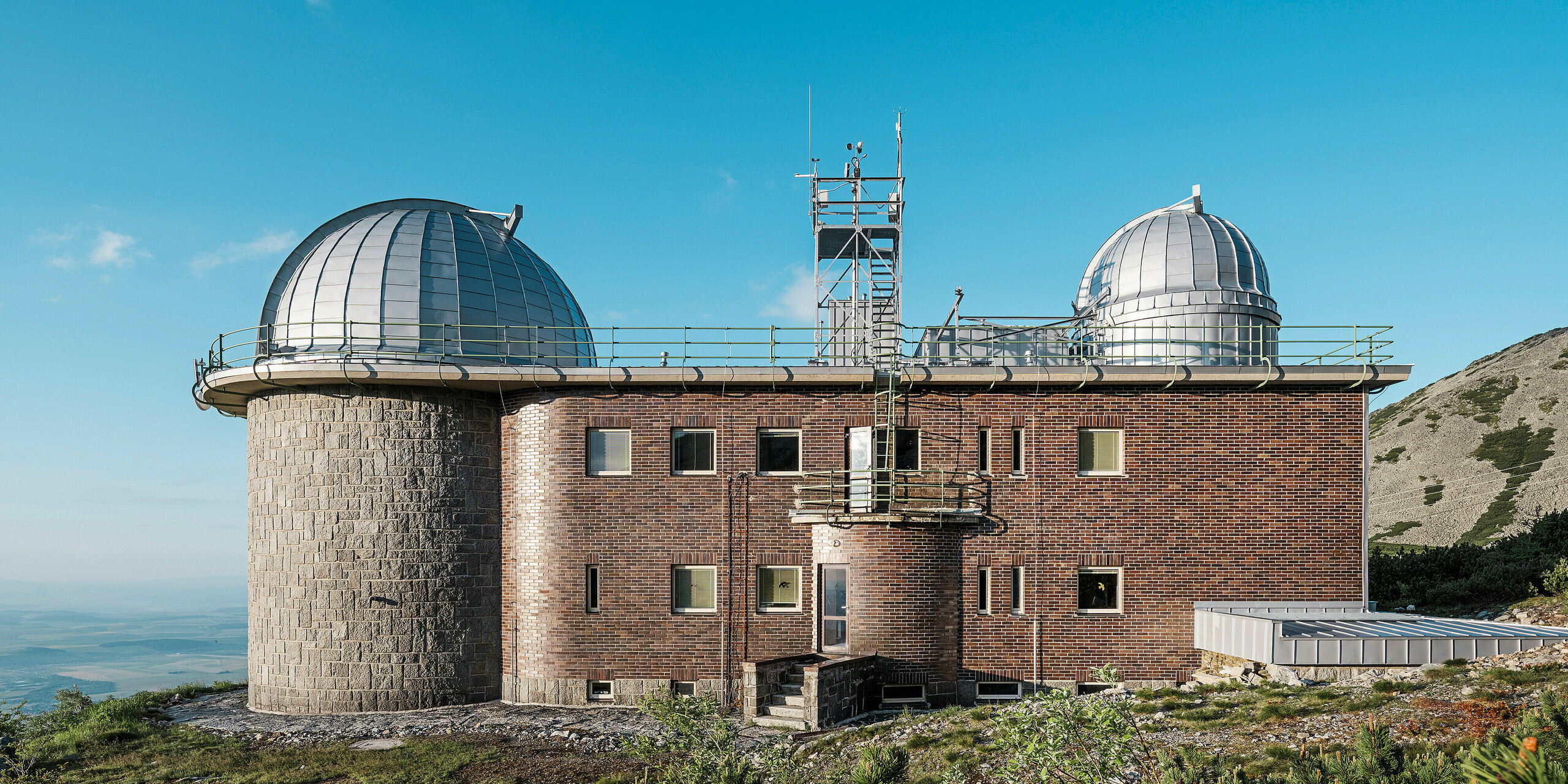 Eine Seitenansicht des Observatoriums von Skalnate Pleso in der Slowakei mit seiner silbermetallisch glänzenden PREFALZ-Kuppel unter einem strahlend blauen Himmel. Die Kombination aus traditioneller Ziegelfassade und modernen Aluminiumelementen steht im Einklang mit der rauen Berglandschaft der Hohen Tatra. Das Gebäude ist ein Beispiel für die harmonische Verbindung von alter Architektur und modernen Baumaterialien. Das denkmalgeschützte Observatorium ist ein leuchtendes Beispiel für Architektur, die sowohl funktional als auch visuell beeindruckt.