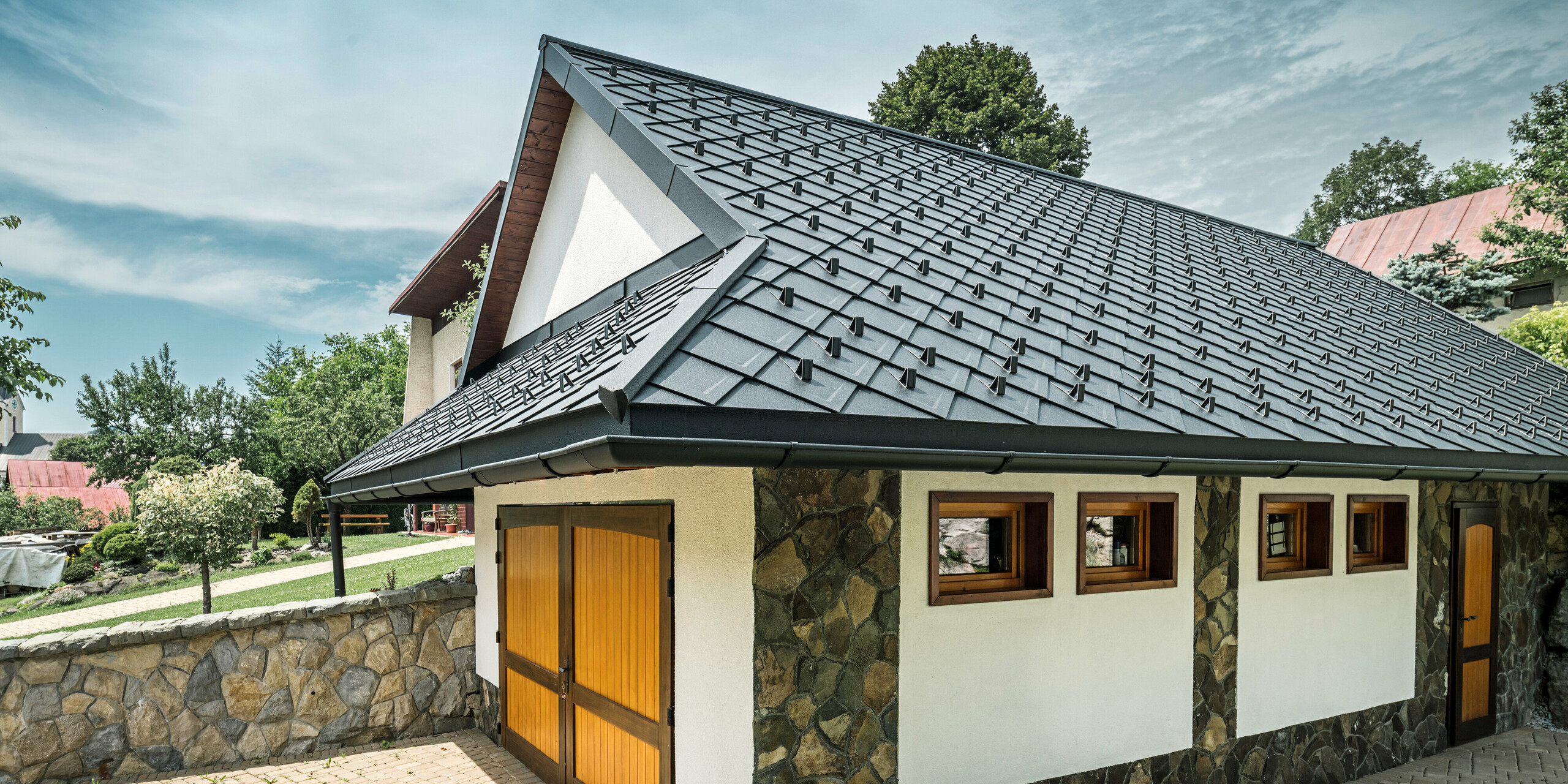 Garage eines traditionellen Einfamilienhauses in Stara Bystrica, Slowakei, mit einem Dach aus PREFA Dachrauten 29 × 29 in P.10 Schwarz. Die robuste Aluminiumdachdeckung in Schuppenoptik harmoniert perfekt mit der Natursteinfassade der Garage. Im Vordergrund ist eine gepflasterte Einfahrt zu sehen, die das gepflegte Erscheinungsbild des gesamten Anwesens unterstreicht.