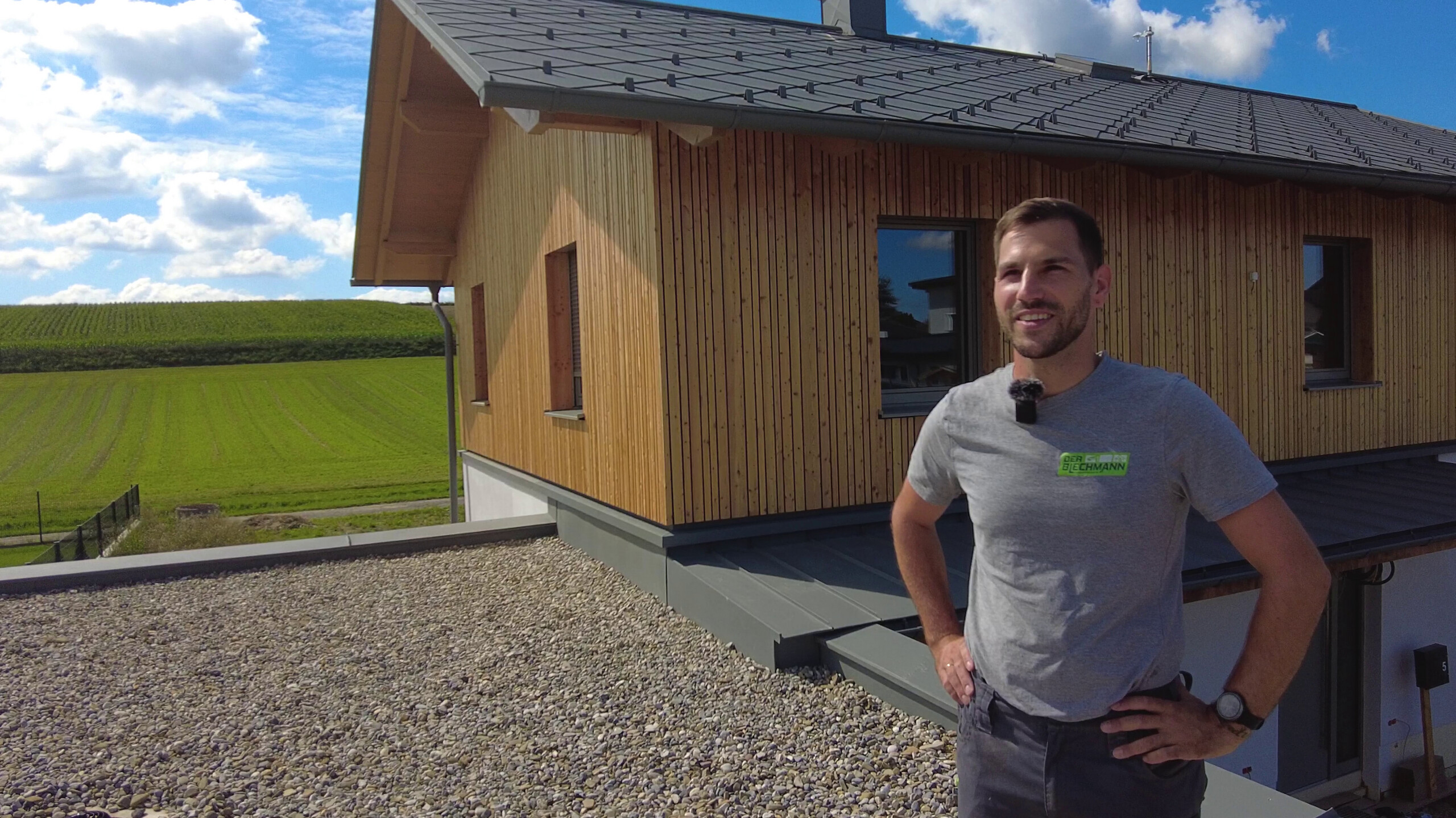 Ein Mann steht auf einer Terrasse eines modernen Einfamilienhauses mit Holzverkleidung. Im Hintergrund sieht man landwirtschaftliche Felder und einen klaren Himmel mit einigen Wolken. Er trägt ein graues T-Shirt mit einem Firmenlogo und lächelt leicht in die Kamera. Das Satteldach des Hauses ist mit PREFA Dachrauten in hellgrau ausgestattet.