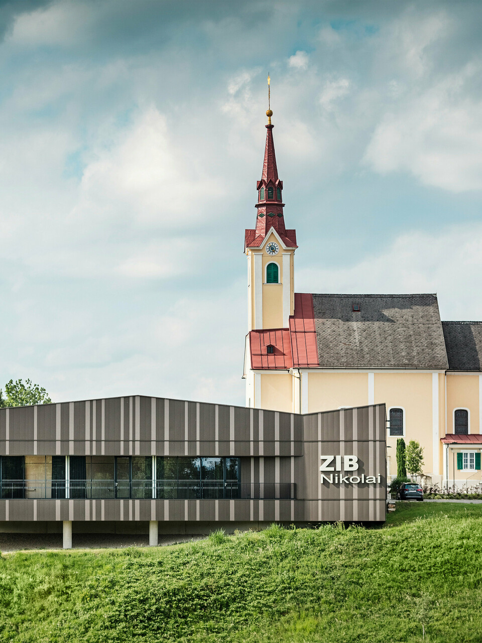 Das Veranstaltungszentrum ZIB Nikolai in St. Veit in der Südsteiermark mit einer modernen, dynamischen Aluminiumfassade in braun-beigegrauen Tönen, im Vordergrund eine grüne Wiese, im Hintergrund die Kirche von St. Nikolai mit gelber Fassade und rotem Dach.