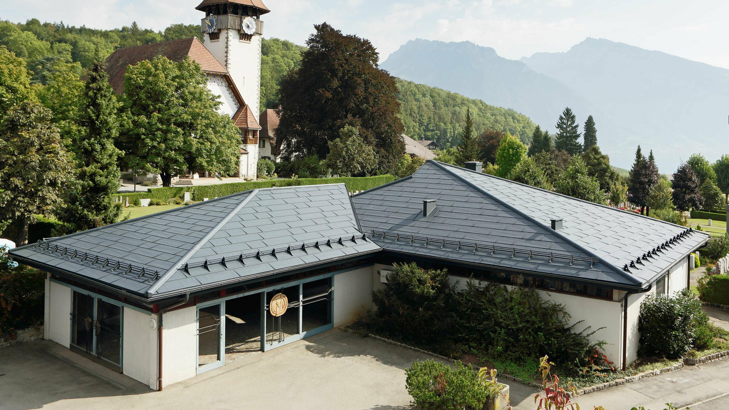 Aufbahrungshalle mit FX.12 Dachpaneelen von PREFA in Anthrazit, im Hintergrund steht eine Kirche und daneben befindet sich der Friedhof.