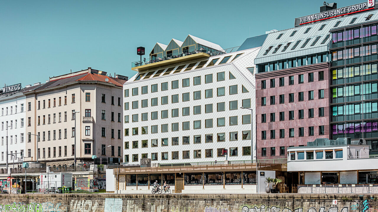 Blick auf das Ufer des Donaukanals in Wien mit einer Reihe von Gebäuden unterschiedlicher Architektur. Im Vordergrund ist das "THE ROCK Radisson RED Vienna" Hotel, erkennbar an seiner charakteristischen weißen PREFA Aluminiumfassade mit Dach- und Wandrauten 44 × 44 in P.10 Reinweiß.