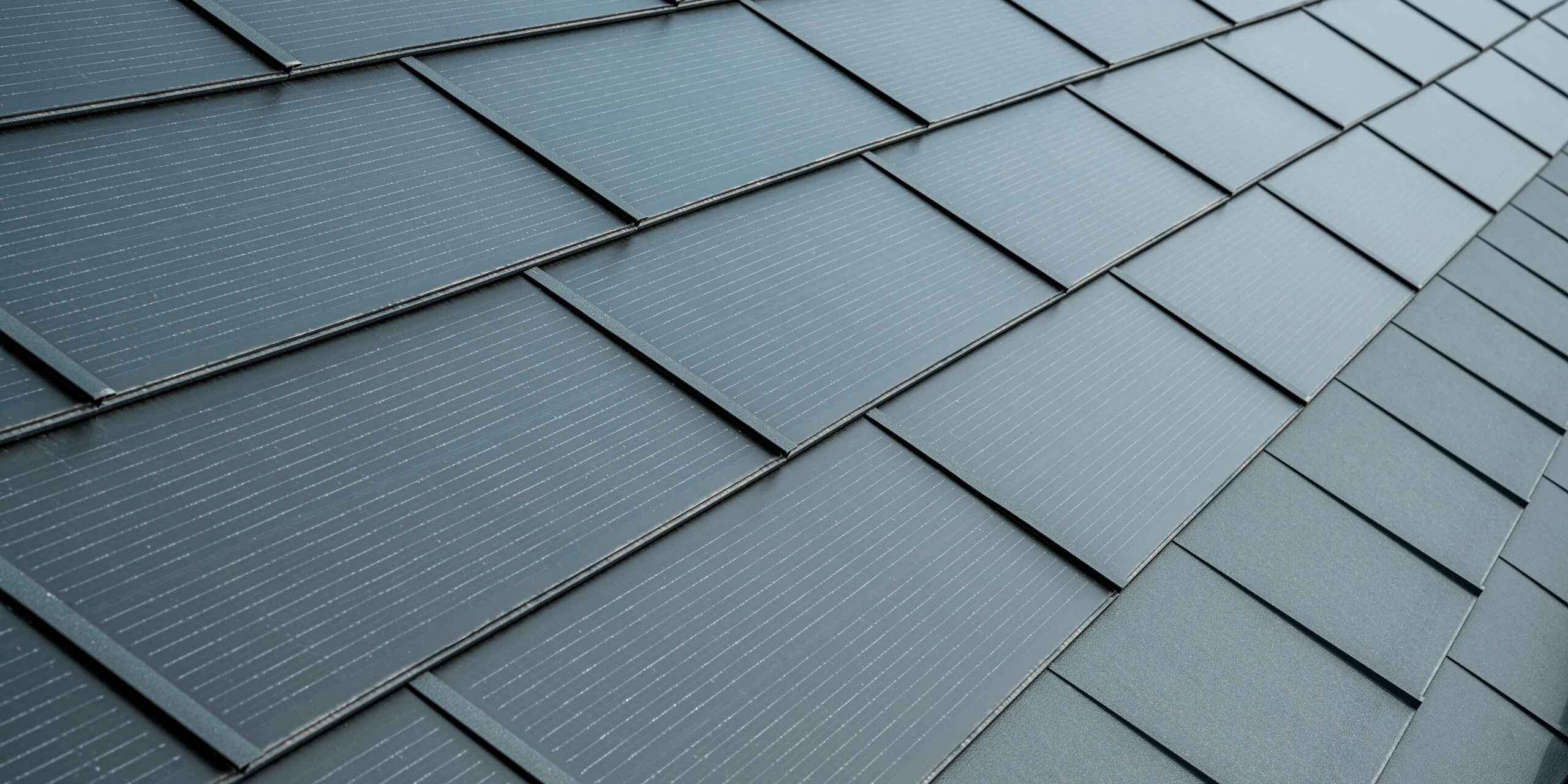 Nahaufnahme eines Daches mit PREFA Solardachplatten in Anthrazit. Die Solarmodule sind elegant in das Dach integriert, wodurch sie sich harmonisch in das Gesamtbild einfügen. Die Dachplatten zeigen eine strukturierte Oberfläche mit feinen Linien, die für zusätzliche Ästhetik sorgen, während sie gleichzeitig als funktionale, energieeffiziente Lösung dienen.