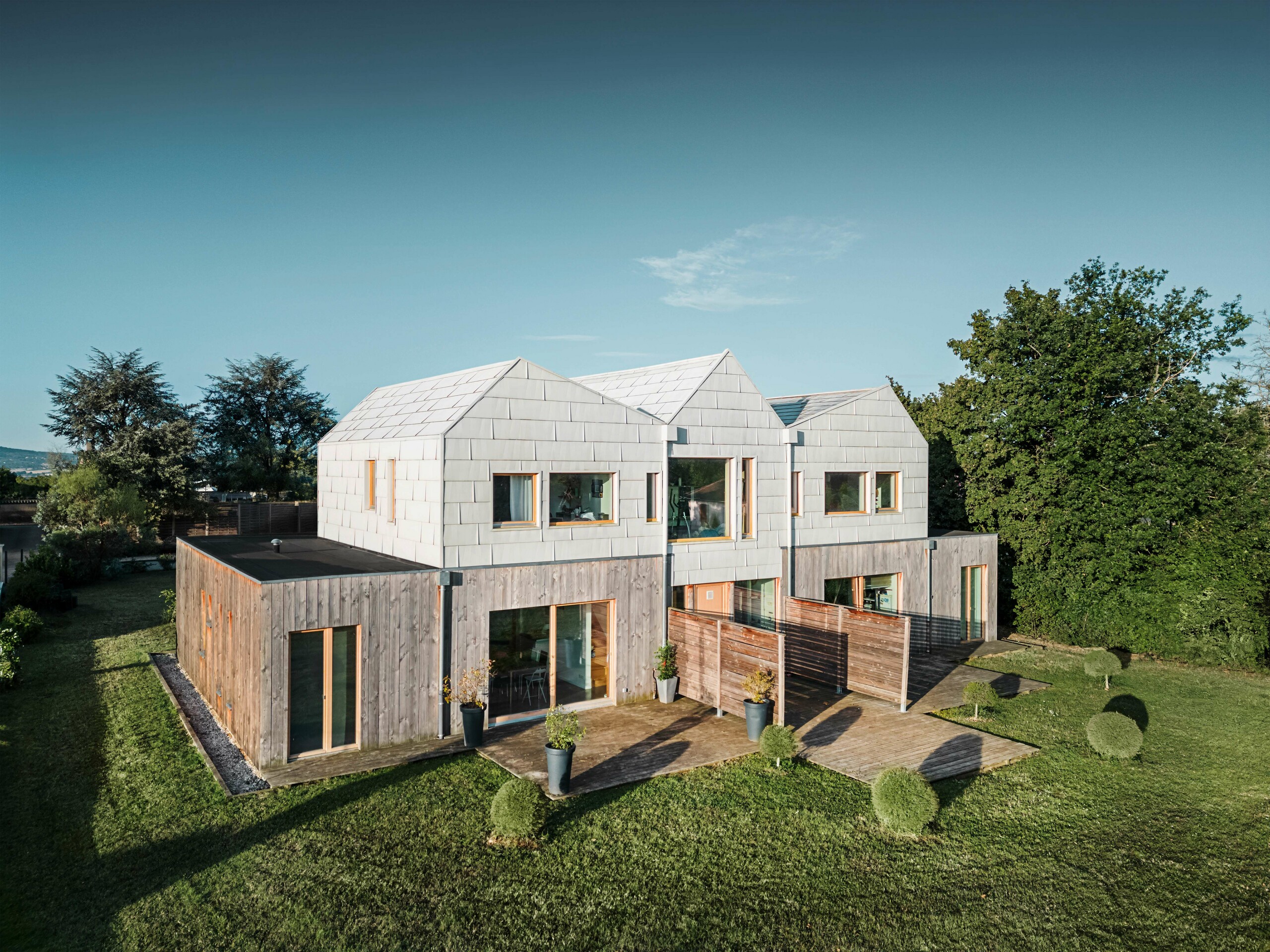 Doppelhaus mit einer Fassade aus FX.12-Aluminiumpaneelen von PREFA, kombiniert mit natürlichen Holzelementen im unteren Bereich. Das Design schafft eine perfekte Harmonie zwischen moderner Architektur und Natur.