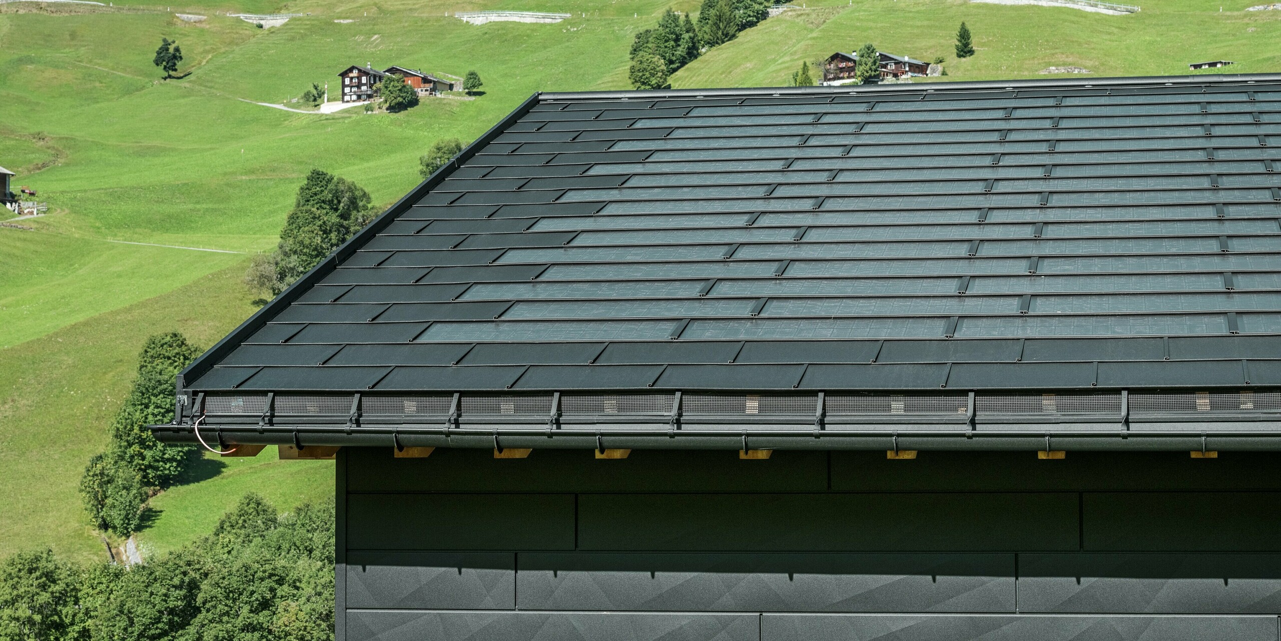 Nahaufnahme der Traufe eines modernen Einfamilienhauses in der alpinen Umgebung von Ascharina in der Schweiz. Das Dach wurde mit der innovativen PREFA Solardachplatte und der traditionellen Dachplatte R.16, beide in P.10 Schwarz, eingedeckt. Für die zuverlässige Dachentwässerung sorgen die PREFA Hängerinne und das PREFA Ablaufrohr. Am unteren Bildrand sind die unverwechselbaren PREFA Siding.X Fassadenpaneele zu sehen. Die klare Linienführung der Aluminiumprodukte unterstreicht das moderne Design inmitten einer malerischen Berglandschaft.