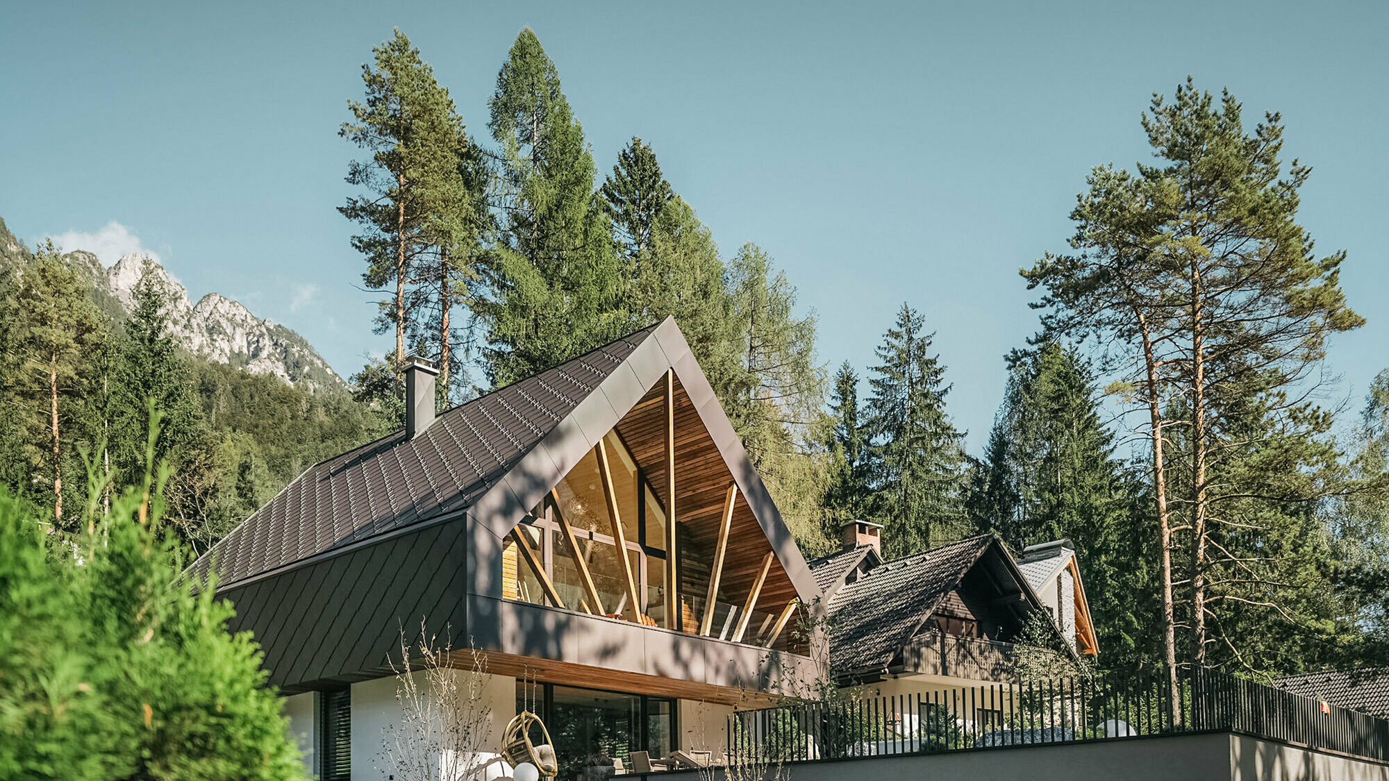 Modernes Alpenchalet in einer idyllischen Berglandschaft, umgeben von hohen Kiefern. Das Haus zeichnet sich durch ein steiles, dunkles Satteldach gedeckt mit der PREFA Dach- und Wandraute 44 × 44 in P.10 Nussbraun und große, dreieckige Fensterfronten aus, die viel Licht in das Innere lassen und den Blick auf die umliegende Natur freigeben. Eine Terrasse mit Geländer erstreckt sich vor dem Erdgeschoss.