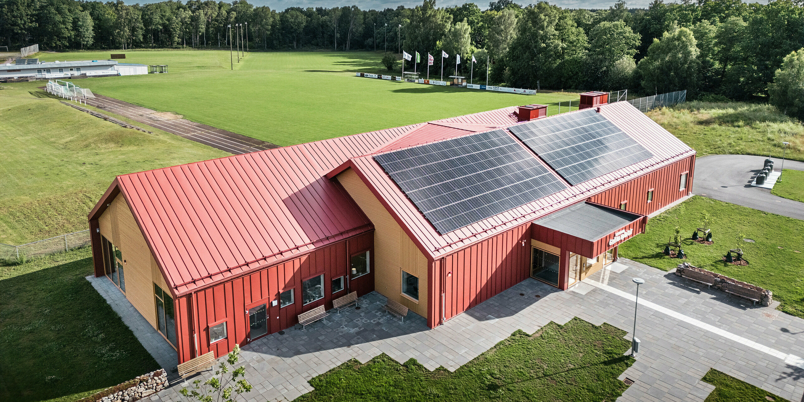 Luftaufnahme der Schulkantine Restaurang Gröna Hjärtat in Broby, Schweden, zeigt das Gebäude mit auffälliger Stehfalzdeckung am Dach und der Fassade aus PREFALZ in Oxydrot. Auf dem Dach ist eine großflächige Photovoltaikanlage installiert, die Strom aus erneuerbarer Energie für das Gebäude liefert. Die klare architektonische Linienführung wird durch die kontrastreiche Kombination aus rotem Aluminium und warmem Holz an den Giebelseiten unterstrichen. Im Hintergrund erstreckt sich ein großes Sportfeld, das in die grüne Umgebung eingebettet ist. Diese Aufnahme betont die harmonische Integration des modernen Gebäudes in die natürliche Landschaft.