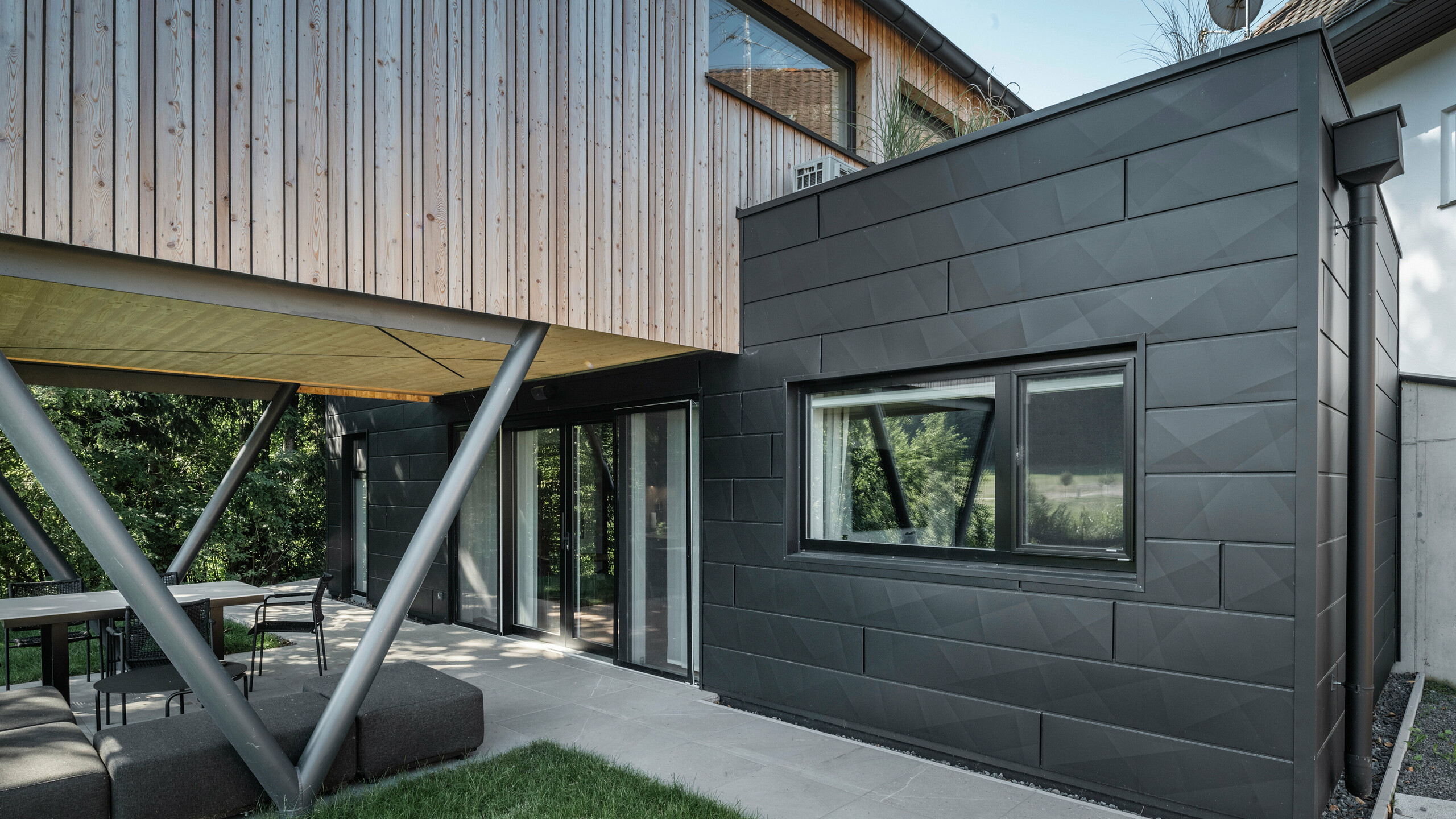 Architektonisches Highlight: 'Woodstock Chalet 5' in St. Martin am Innkreis, Oberösterreich. Die moderne Unterkunft für Hochzeiten wird durch die Verwendung von anthrazitfarbenen PREFA Siding.X-Fassadenpaneelen unterstrichen. Die Aluminium-Elemente bilden einen starken Kontrast zu den hellen Holzpaneelen und den metallischen Stützstrukturen. Die dunklen Fensterrahmen fügen sich harmonisch in die Fassadengestaltung ein, wodurch das Gebäude eine elegante Ausstrahlung erhält. Dieses Bild zeigt die ästhetische Balance zwischen natürlicher Holztextur und dem industriellen Charme der Aluminiumfassade, die für die Gestaltung zeitgemäßer Gebäude steht.