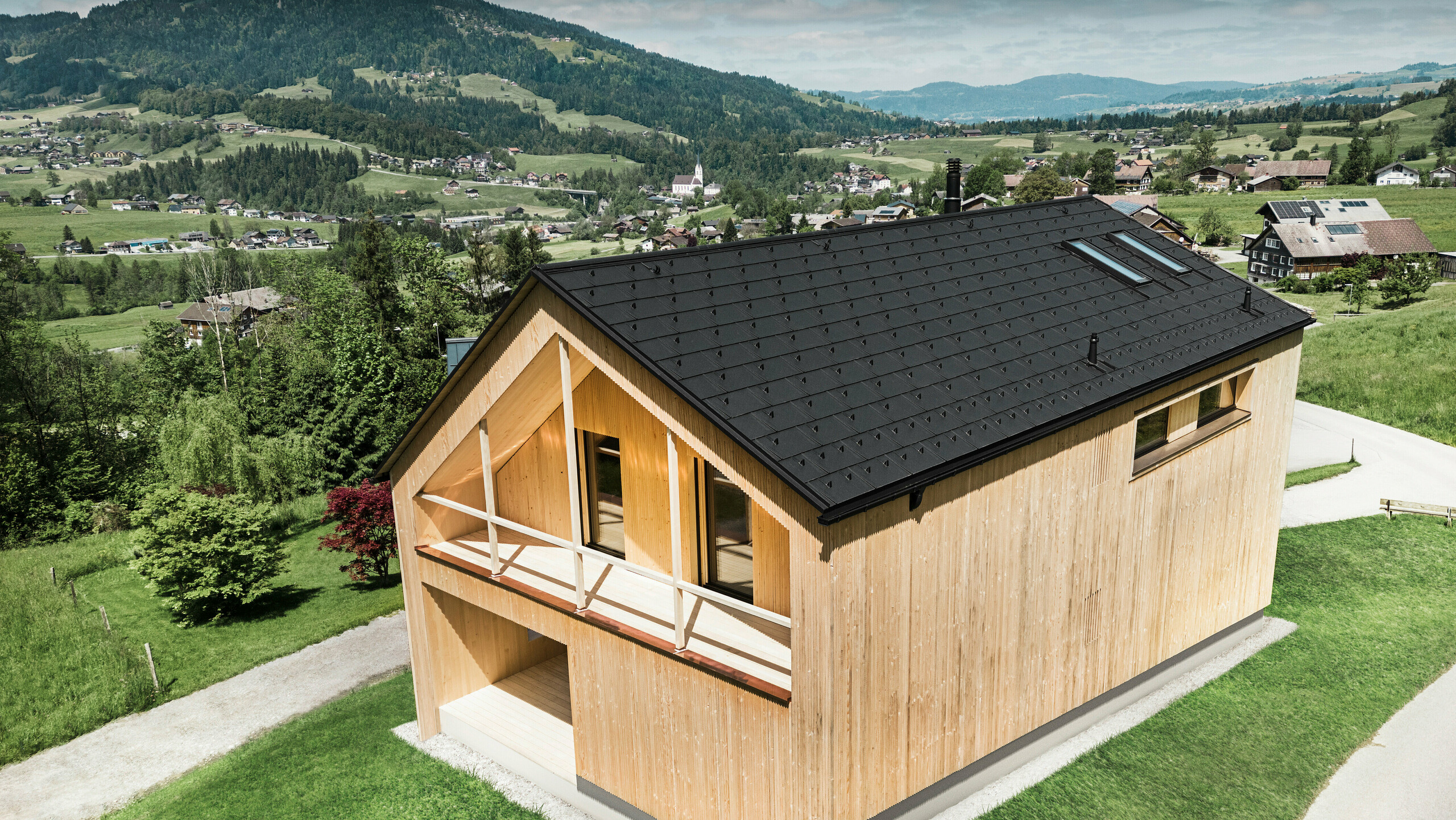 Dieses Foto zeigt ein ökologisches Vorzeigehaus in Egg, das sich mit seiner PREFA Solardachplatte und den schwarzen Dachplatten R.16 gegen die malerische Kulisse eines österreichischen Dorfes abhebt. Die Solarelemente sind nahtlos in die Dachstruktur eingebettet und liefern Energie aus einer erneuerbaren Energiequelle, während das natürliche Holz der Fassade und die sattgrüne Umgebung eine Verbindung von Nachhaltigkeit und Lokalität darstellen. Der Blick auf die umliegende dörfliche Struktur mit traditionellen Bauten im Hintergrund verleiht dem Bild eine zusätzliche Dimension und betont die gelungene Integration moderner, umweltfreundlicher Bauweisen in gewachsene Landschaften.