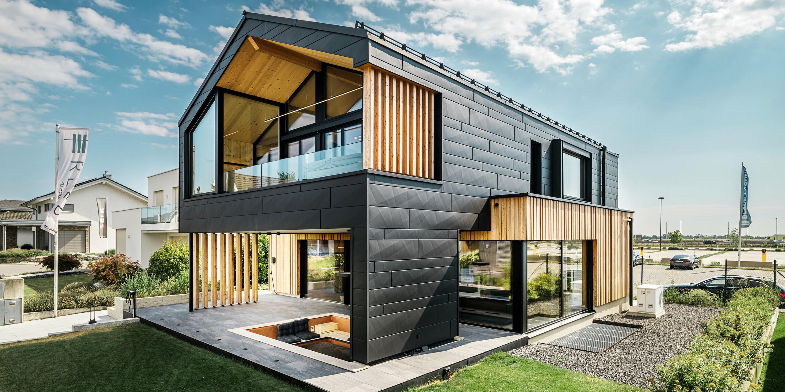 Das Musterhaus Autark im Fertighauszentrum Blaue Lagune in Vösendorf (AT) zeigt eine moderne Kombination aus extravaganten PREFA Siding.X Fassadenpaneelen in P.10 schwarz und natürlichen Holzelementen. Die große Loggia und der Balkon fügen sich harmonisch in die zeitgemäße Gestaltung ein. Die Holzelemente schaffen eine einladende Atmosphäre und die robuste Aluminiumfassade sorgt für Langlebigkeit und Witterungsbeständigkeit. Die großzügige Gartenanlage mit Terrasse und eingebautem Sitzplatz rundet das außergewöhnliche und nachhaltige Architekturkonzept ab.