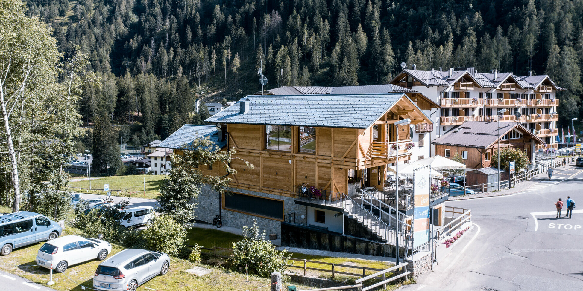 Luftaufnahme eines alpinen Après-Ski-Lokals in Italien, hervorgehoben durch ein widerstandsfähiges Dachsystem aus Aluminium. Die robusten PREFA Dachschindeln DS.19 in P.10 Steingrau schützen das Gebäude vor Unwetter. Die kontrastierende Kombination aus Holzstruktur und steingrauen Akzenten, umgeben von dichten Kiefernwäldern und traditioneller Chalet-Architektur, schafft ein einladendes Ambiente für Besucher und integriert sich harmonisch in die naturbelassene Umgebung.