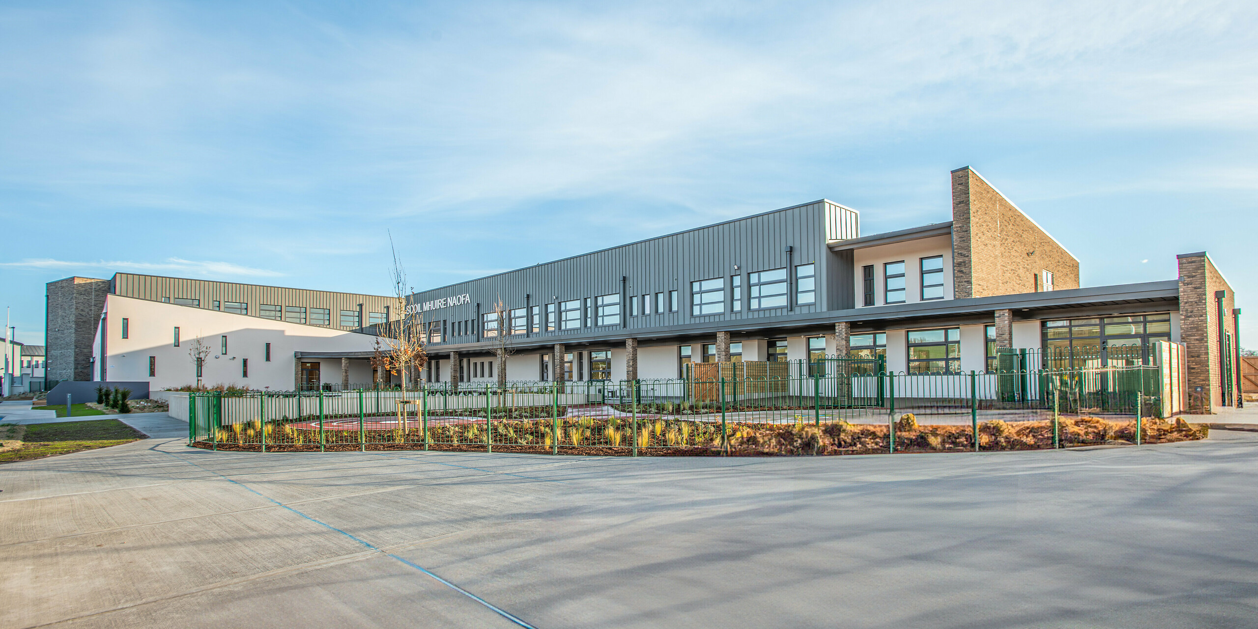 Weitwinkelperspektive der Scoil Mhuire Naofa in Carrigtwohill, Irland. Das Schulgebäude verbindet moderne Architektur mit einer PREFALZ Aluminiumfassade in P.10 Hellgrau. Die langlebige und witterungsbeständige Stehfalzdeckung unterstreicht die funktionale Eleganz, während die gepflegten Außenanlagen den modernen Charakter der Schule unterstreichen.