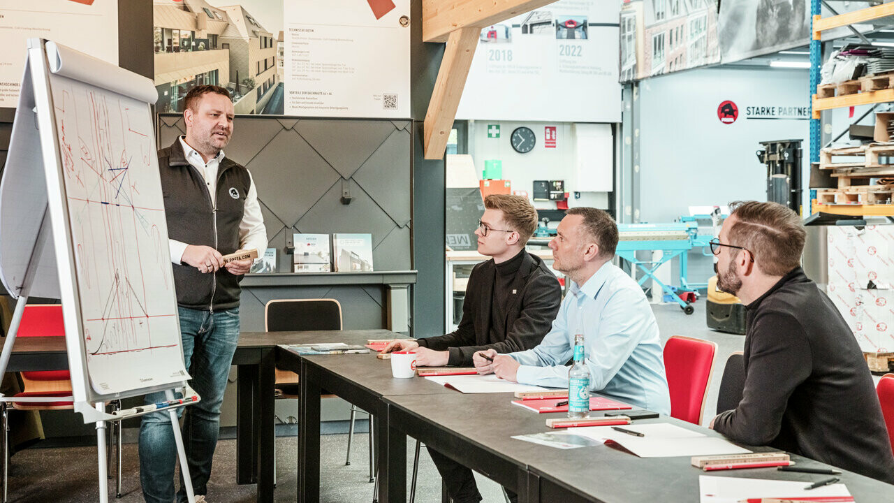 Ein Mitarbeiter der PREFA Academy steht vor einem Flipchart und erklärt technische Details zu Dachmaterialien. Drei Teilnehmer sitzen an einem Tisch und folgen aufmerksam der Präsentation. Im Hintergrund sind Informationsposter und technische Geräte zu sehen, die den praxisorientierten Ansatz der Schulung unterstreichen.