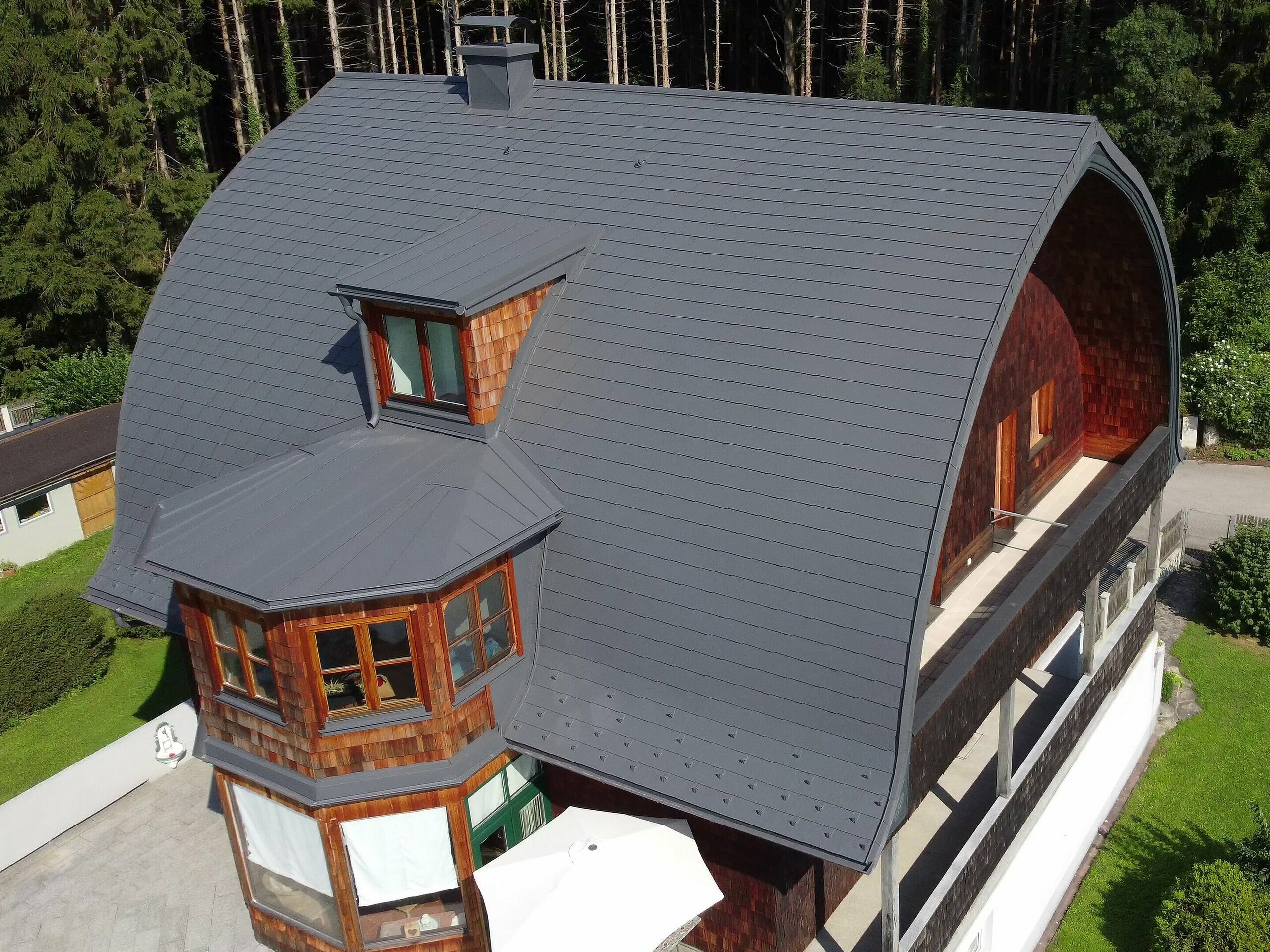 Einfamilienhaus mit geschwungenem Tonnendach, gedeckt mit PREFA Dachschindeln in P.10 Dunkelgrau nach einer umfassenden Dachsanierung. Das Haus ist von einem gepflegten Garten umgeben, mit einem großen, hellen Terrassenbereich und Sonnenschirm im Vordergrund. Im Hintergrund sieht man dichte Nadelwälder, die das Grundstück umrahmen, sowie eine ruhige, angrenzende Straße.