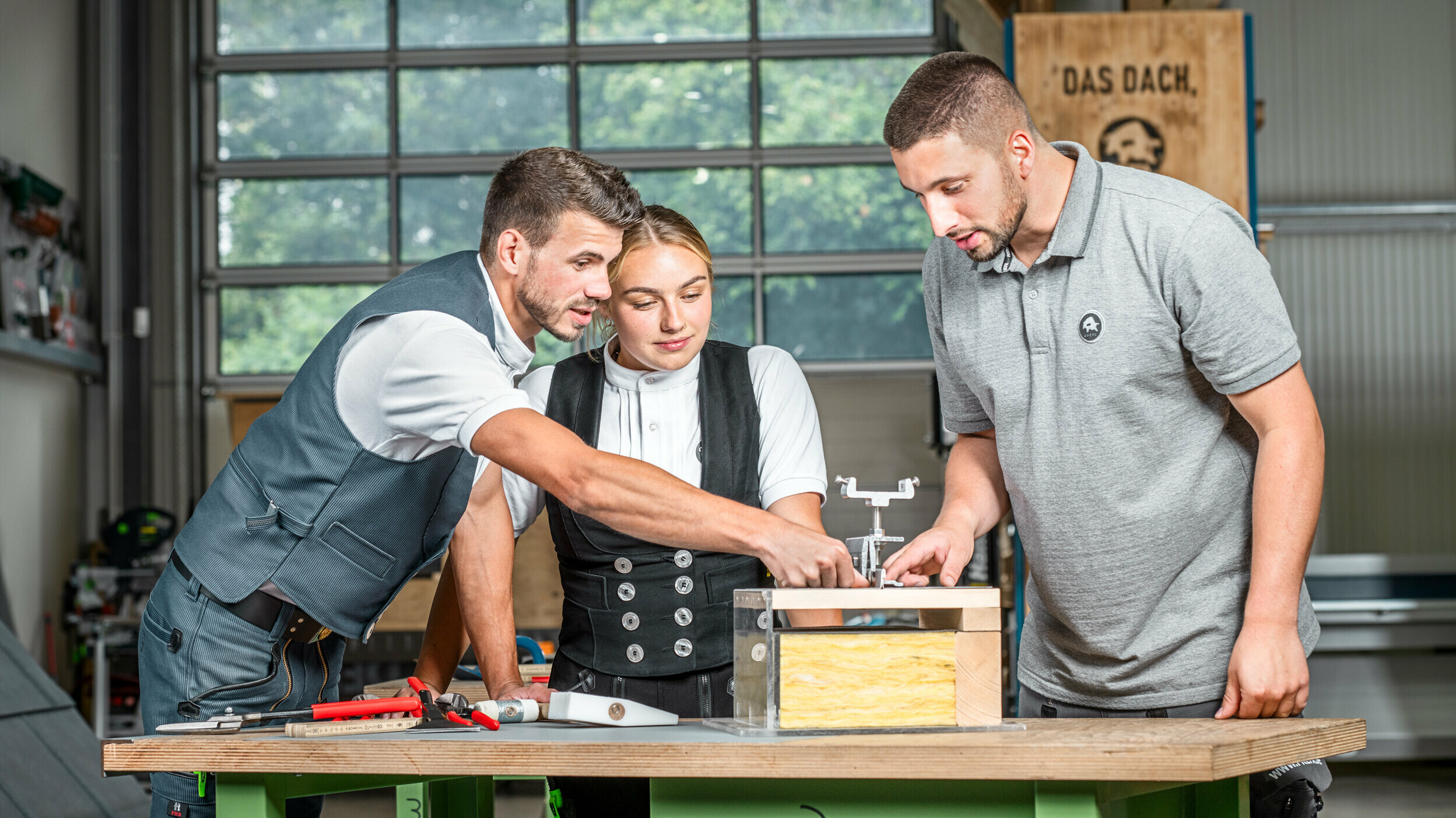 PREFA Anwendungstechniker Julian Eickhoff, Marvin Bürger von Bürger Falztechnik und Dachdeckermeisterin Fabienne Ellermeier schauen sich gemeinsam ein Lehrmodell in der PREFA Academy Rheinbach an.