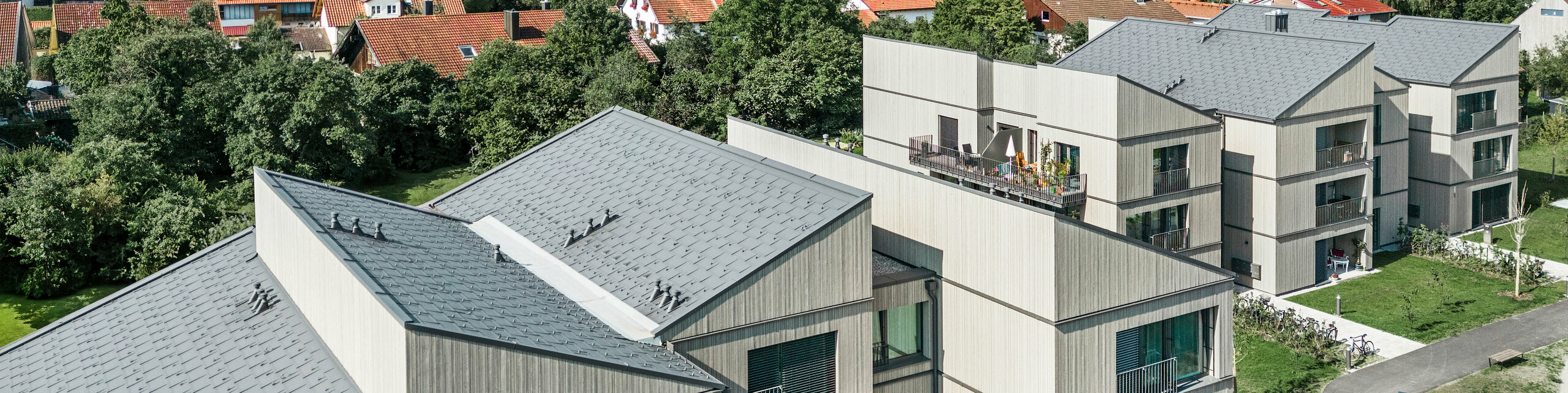 Drohnenaufnahme des modernen Gebäudekomplexes am Schmucker-Areal in Utting am Ammersee (Deutschland), mit innovativer Holzhybridbauweise und langlebigen PREFA Dachplatten R.16 in P.10 Dunkelgrau. Die Gebäude zeichnen sich durch ihre klaren Linien und nachhaltige Architektur aus, kombiniert mit eleganten Holzfassaden und großzügigen Balkonen. Die hochwertige Verarbeitung der PREFA Aluminiumprodukte sorgt für optimale Wetterbeständigkeit und Langlebigkeit, während die harmonische Integration in die grüne Umgebung eine einladende Wohnatmosphäre schafft. Im Vordergrund ist ein begrünter Spielplatz zu sehen, der zur familienfreundlichen Gestaltung des Areals beiträgt.