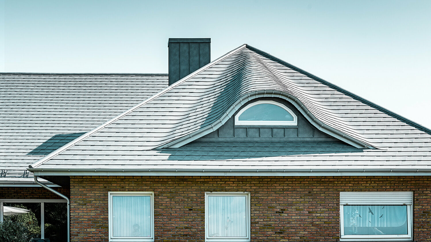 Einfamilienhaus aus den 70ern mit einem Schindeldach aus Aluminium in P.10 Steingrau mit geschwungener Gaube mit rundem Fenster