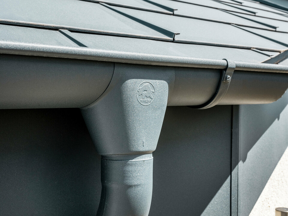 PREFA Hängerinne mit Rinnenkessel in P.10 Anthrazit und der PREFA Dachplatte R.16 Nahaufnahme eines Hausdaches mit PREFA Dachplatte R.16 in Anthrazit sowie detailreicher Ansicht der farblich abgestimmten PREFA Dachrinne und Fallrohr aus Aluminium bei sonnigem Wetter.