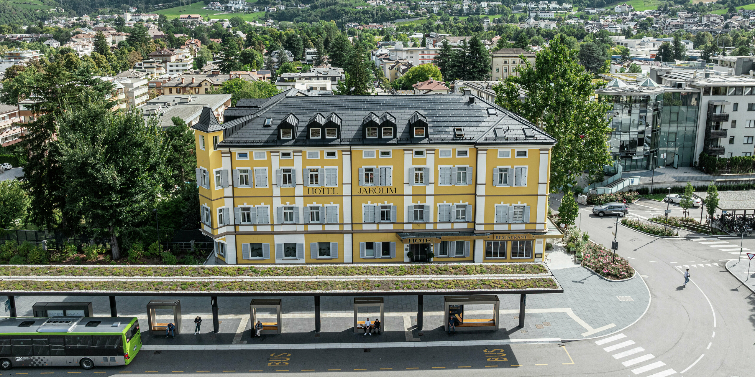 Blick von oben auf das renovierte Hotel Jarolim am Bahnhof in Brixen mit einem Dach aus PREFA Solardachplatten und Dachplatten R.16. Das robuste Aluminiumdach überzeugt durch seine langlebige, witterungsbeständige und leichte Konstruktion, ideal für denkmalgeschützte Gebäude. Die harmonisch integrierten Photovoltaikmodule sorgen für eine nachhaltige Energiegewinnung, ohne die historische Ästhetik zu beeinträchtigen. Der markante Turm wurde mit robusten PREFA Dachrauten gestaltet.