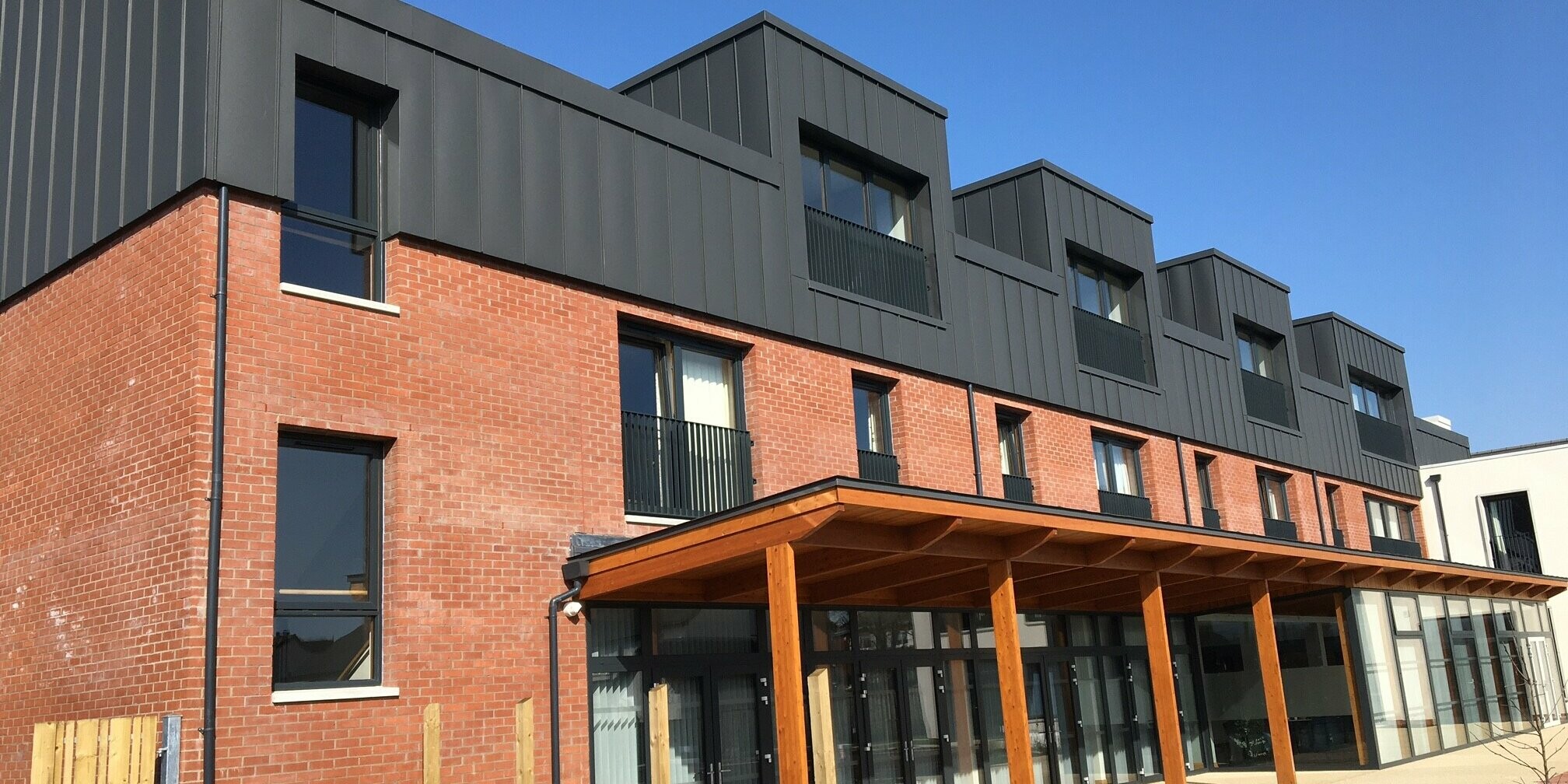 Blick vom Garten auf die Harberton Hall in Belfast, Nordirland. Die Stehfalzdeckung aus dunkelgrauem PREFALZ Aluminium trifft auf die Ziegelfassade. Ein begrünter Außenbereich und Flachdachgauben verstärken die harmonische Verbindung von moderner Architektur und traditionellem Stil in dieser betreuten Wohnanlage.