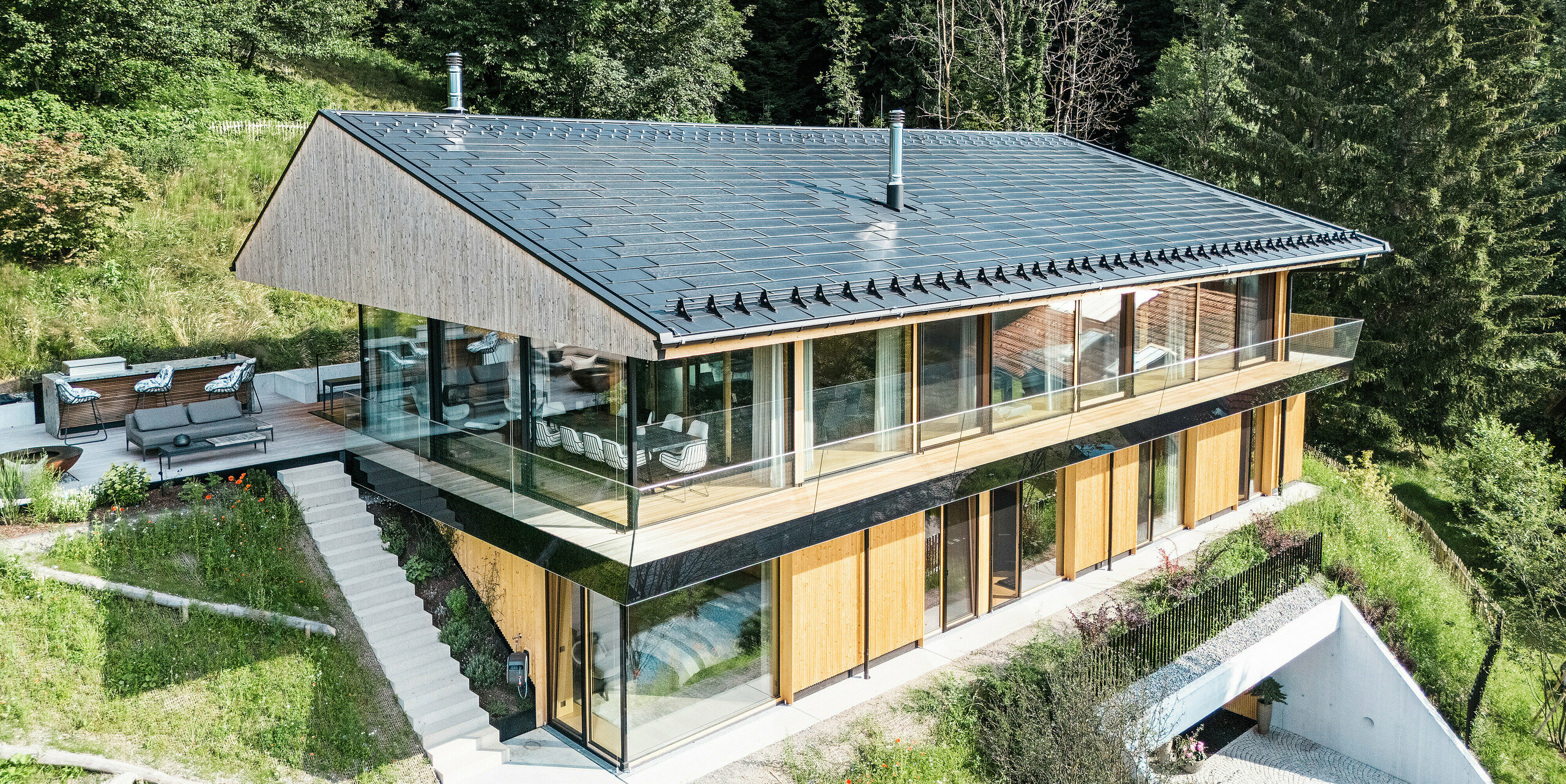 Vogelperspektive auf ein modernes Einfamilienhaus in Egg, Vorarlberg, mit PREFA Solardachplatten und Dachplatten R.16 in P.10 Schwarz. Die Dachkonstruktion aus Aluminium sorgt für nachhaltige Energiegewinnung und bietet robusten Schutz vor Witterungseinflüssen. Der Schneeschutz verhindert in den Wintermonaten abrutschende Dachlawinen. Die großzügige Glasfassade und die Holzverkleidung runden das formschöne Design ab, während im Hintergrund die Terrasse mit hochwertigen Gartenmöbeln zu sehen ist.