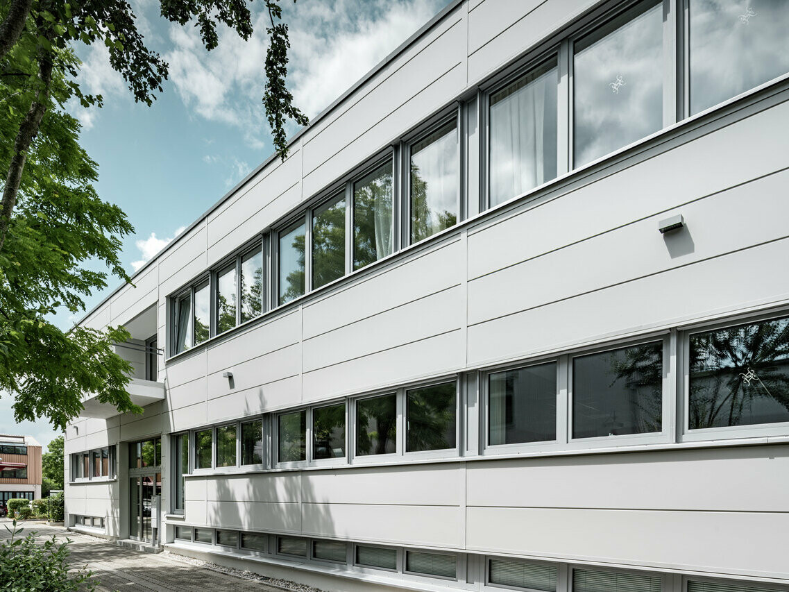 Seitenansicht der PREFA Sidings in der Farbe Prefaweiss, das Objekt hat mehrere Fenster, wobei die Wolken des Himmels gespiegelt werden.