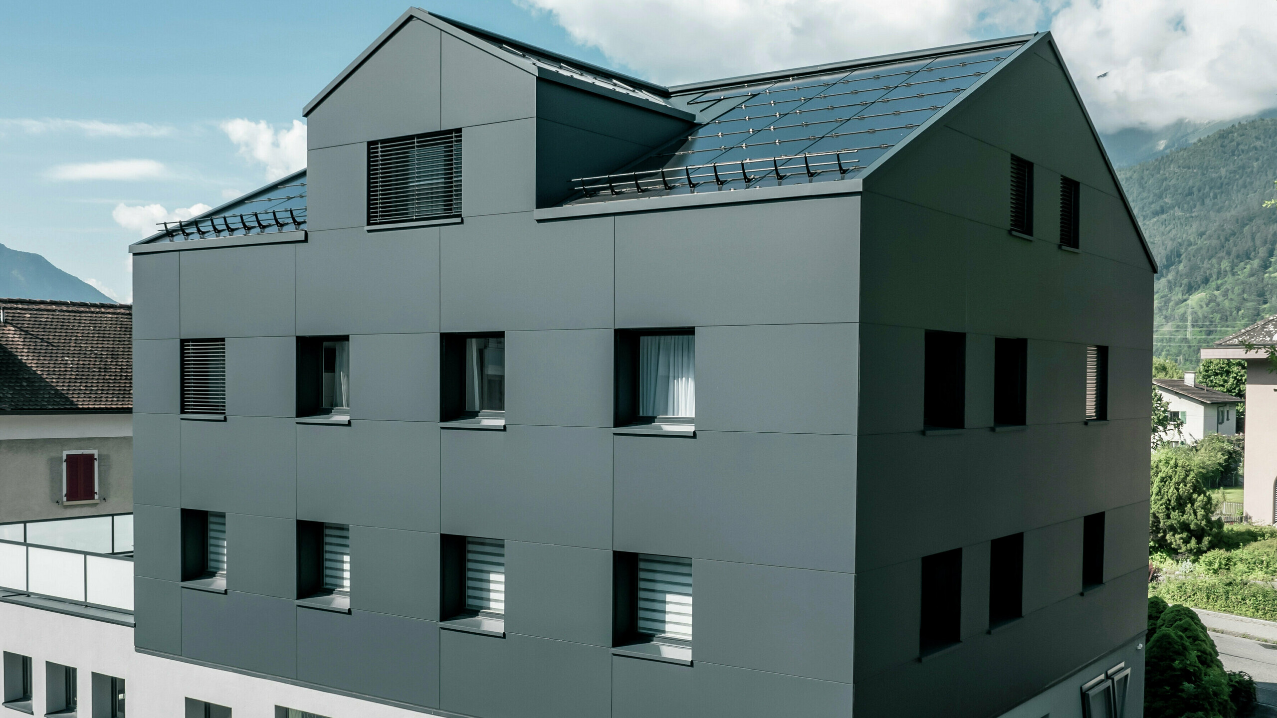 Modernisiertes Wohn- und Geschäftsgebäude nach der Fassadensanierung mit PREFABOND Aluminium Verbundplatten in Anthrazit. Die klare, elegante Linienführung der neuen Fassade verleiht dem Gebäude ein modernes Erscheinungsbild. Im Erdgeschoss befindet sich das Architekturbüro Voser. Die Umgebung mit Bäumen und Bergen rundet die harmonische Optik ab.