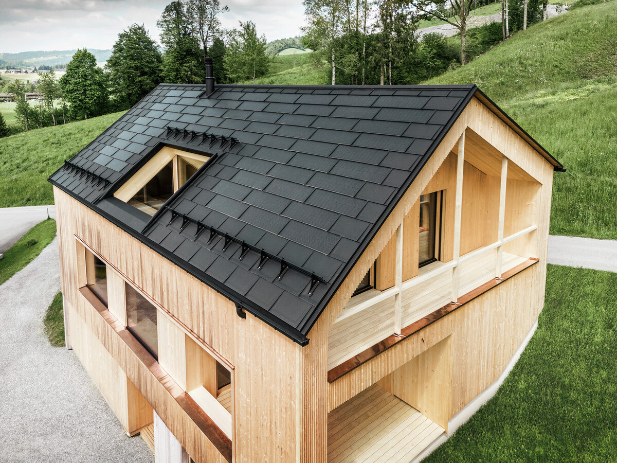 Vogelperspektive auf ein umweltfreundliches Holzhaus in Egg, eingebettet in grüne Wiesen, mit einem dunklen, langlebigen PREFA Solardach. Die schwarzen Dachplatten R.16 aus Aluminium harmonieren mit den Solarmodulen, die effizient in das Dach des Einfamilienhauses integriert sind, um nachhaltige Energie zu erzeugen. Das Gebäude mit seiner charakteristischen Holzfassade und modernen Architektur strahlt Natürlichkeit und zukunftsorientiertes Design aus, während es die Landschaft mit seiner fortschrittlichen Solartechnologie respektiert.