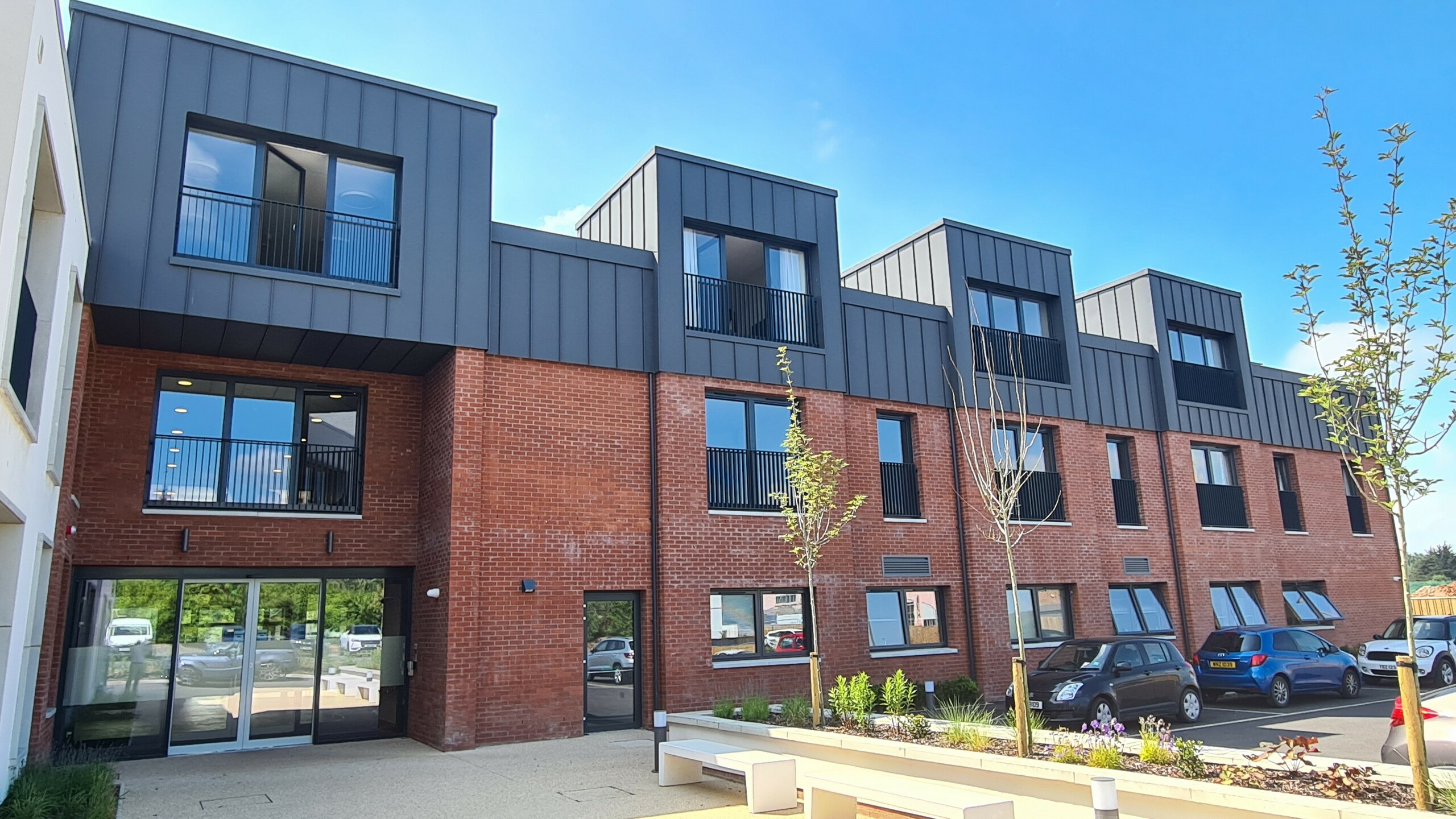 Vorderansicht der Harberton Hall in Belfast, Nordirland. Die Stehfalzdeckung aus PREFALZ P.10 dunkelgrau ergänzt die Ziegelfassade perfekt. Große Fenster und Flachdachgauben unterstreichen das innovative Design des Gebäudes für betreutes Wohnen und schaffen eine einladende Atmosphäre. Die gläserne Eingangstür gewährt Einblicke in das Innere der Einrichtung.