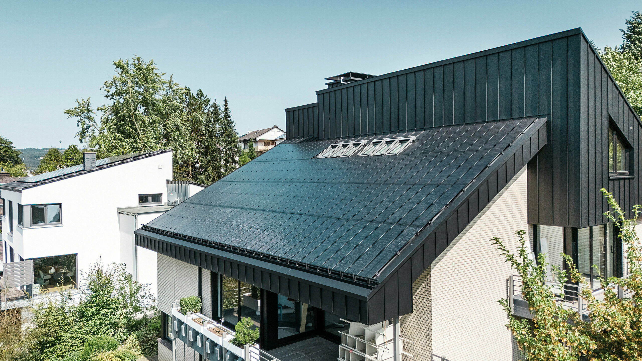 Moderne Architektur eines Einfamilienhauses in Meschede, Deutschland, mit einem PREFALZ Aluminiumdach in P.10 Schwarz und integrierten Glas/Glas PV-Modulen. Das versetzte Pultdach verbindet zeitloses Design mit nachhaltiger Photovoltaik-Technologie. Die innovativen PREFALZ Solarmodule liefern eine Leistung von 18 kWp. Die rahmenlosen Module und die elegante Aluminiumverkleidung von Fassade und Dach bilden eine ästhetische und funktionale Einheit, die sich perfekt in das grüne Wohnumfeld einfügt.